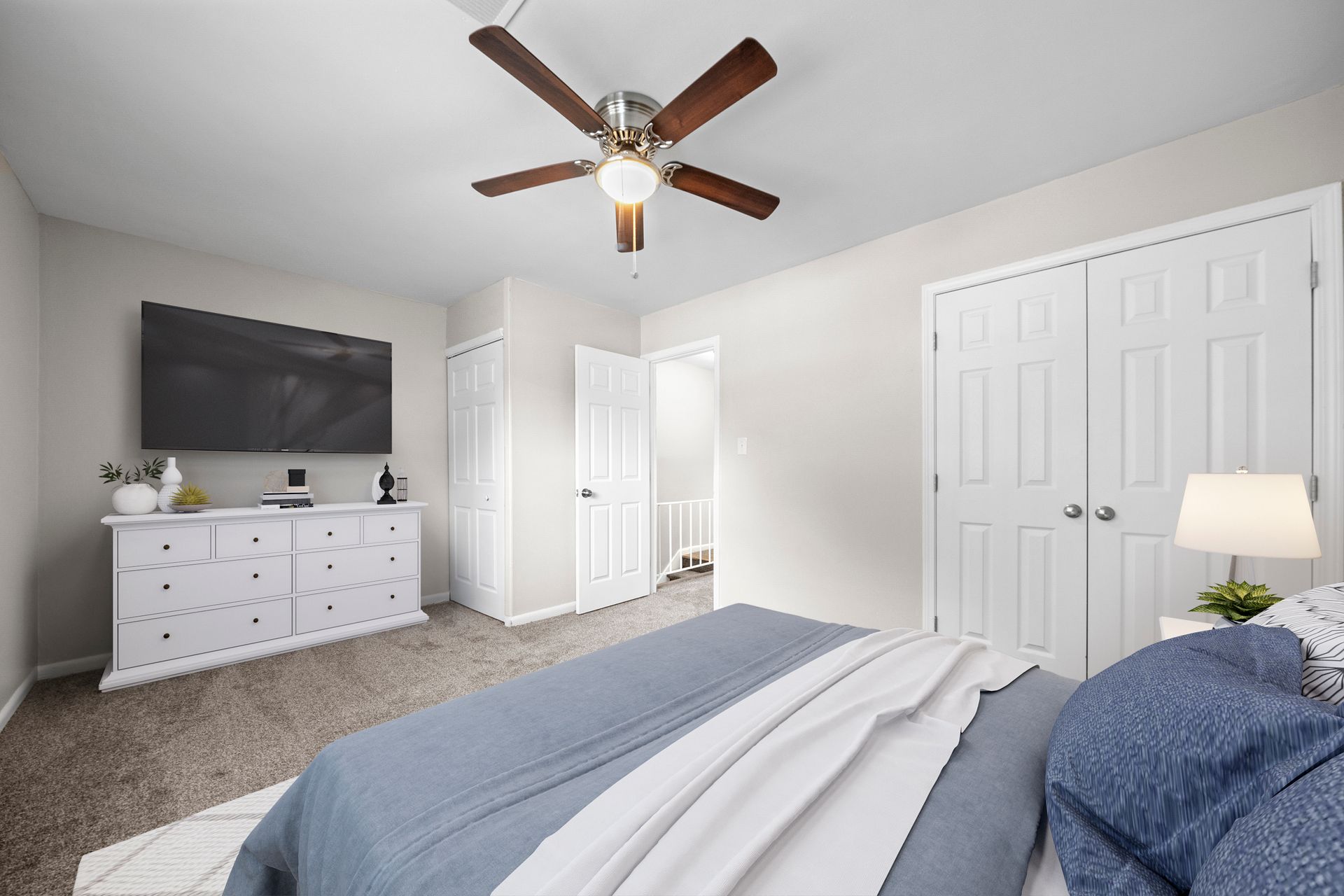 Bright bedroom with a bed, dresser, ceiling fan, white closet doors, and a wall-mounted TV.