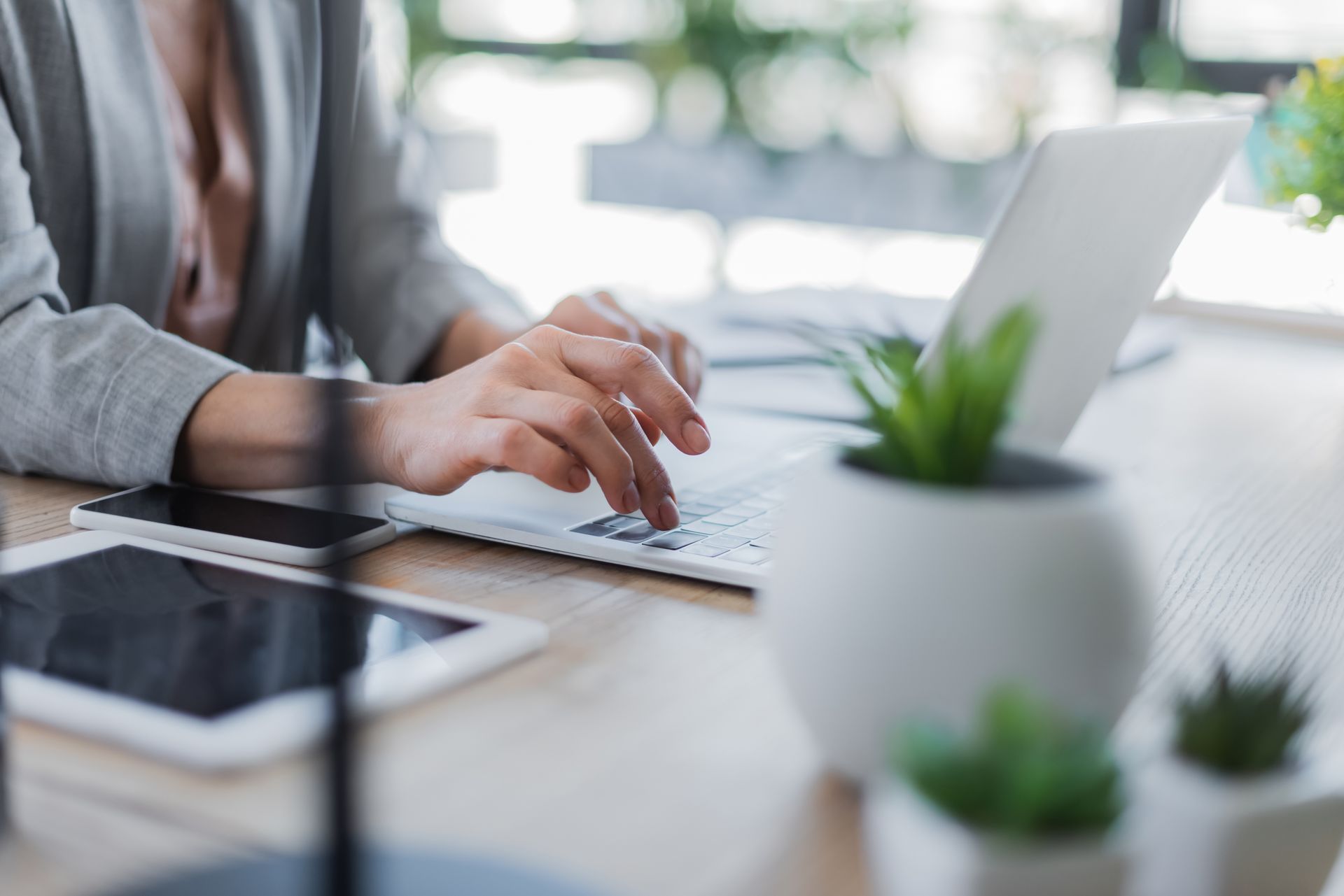 Hands typing on a laptop at a bright desk with a tablet and small potted plants nearby