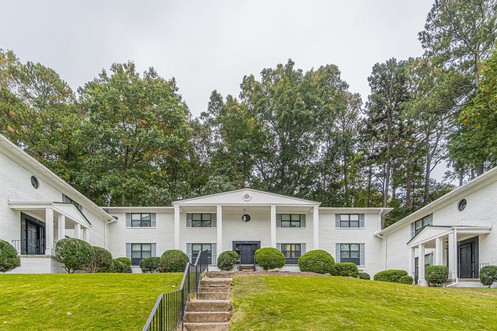 White apartment building with central stairs, green hedges, and trees in front