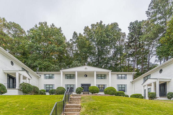 White apartment building with central stairs, green hedges, and trees in front
