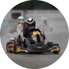 Go-kart racer in black and orange suit speeding on a wet track with blurred competitors behind