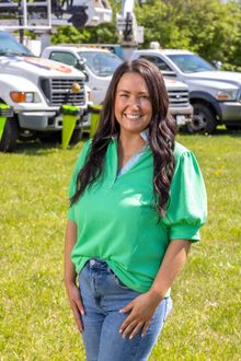 Woman in a green shirt and jeans smiling outdoors on a grassy lawn with parked cars behind her