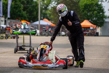 Kart driver in black suit stands beside a crashed red-and-white kart on a track.
