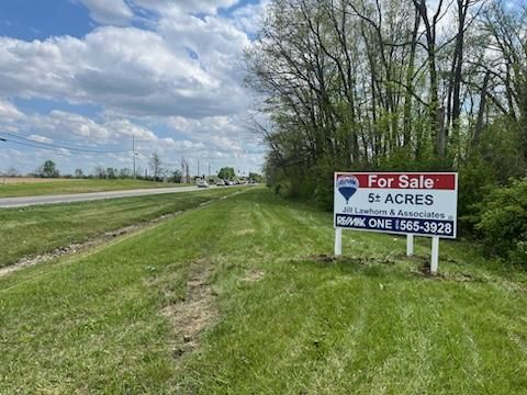 For Sale sign on vacant roadside acreage beside grassy ditch and trees under cloudy sky