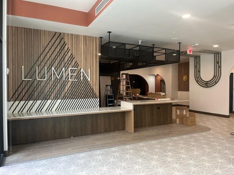 Lobby reception desk with LUMEN sign, wood slat wall, and patterned carpet