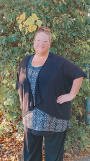 Person standing outdoors in front of leafy greenery, wearing a black cardigan and patterned top.