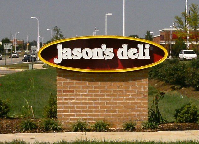Jason's Deli sign on a brick base beside a roadside, with grass and buildings in the background