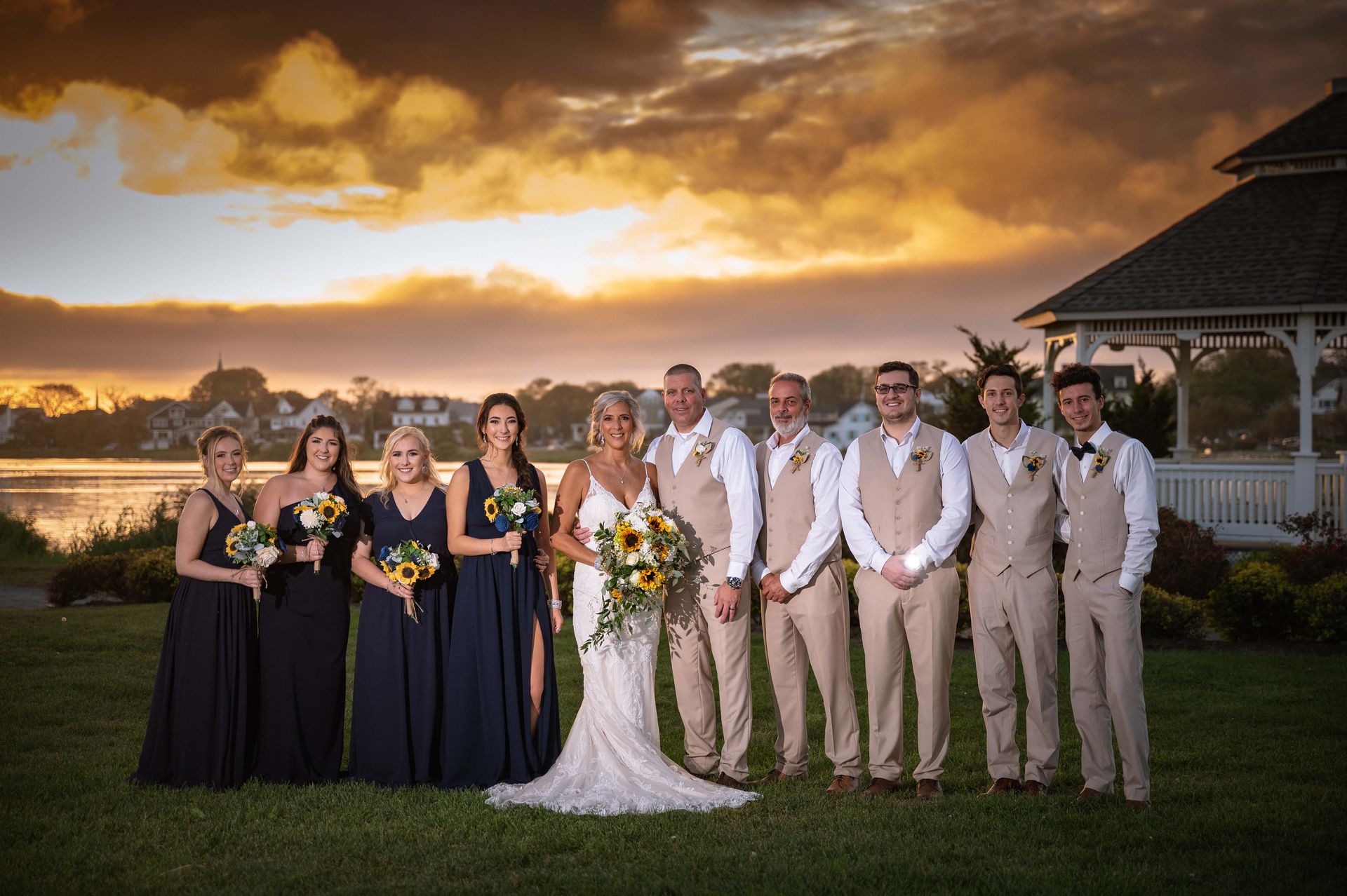 Wedding party posing in front of lake and gazebo at sunset, with a bride in white dress and attendants in navy and tan attire.