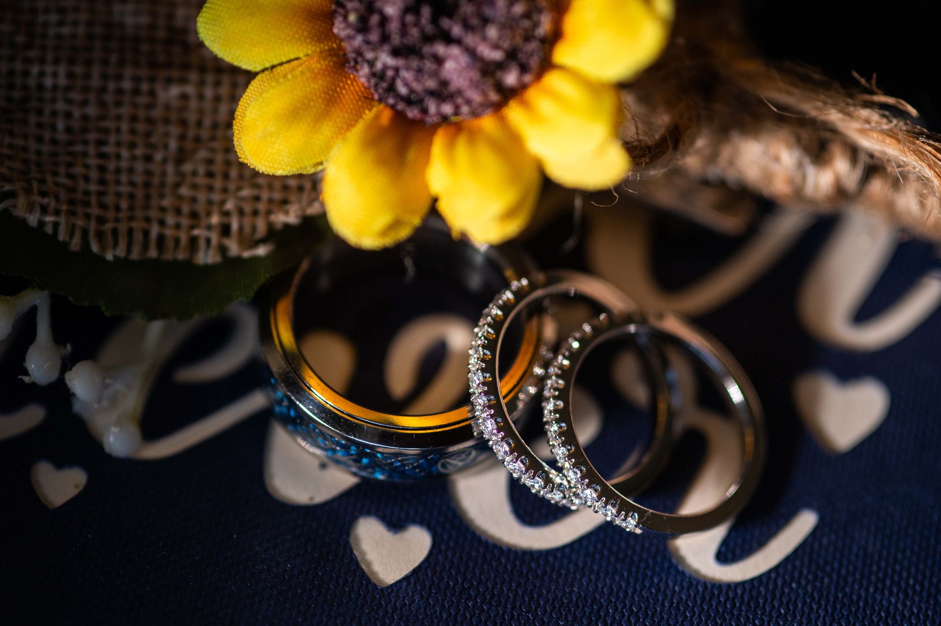Wedding rings on blue surface with carved hearts and sunflower in background.