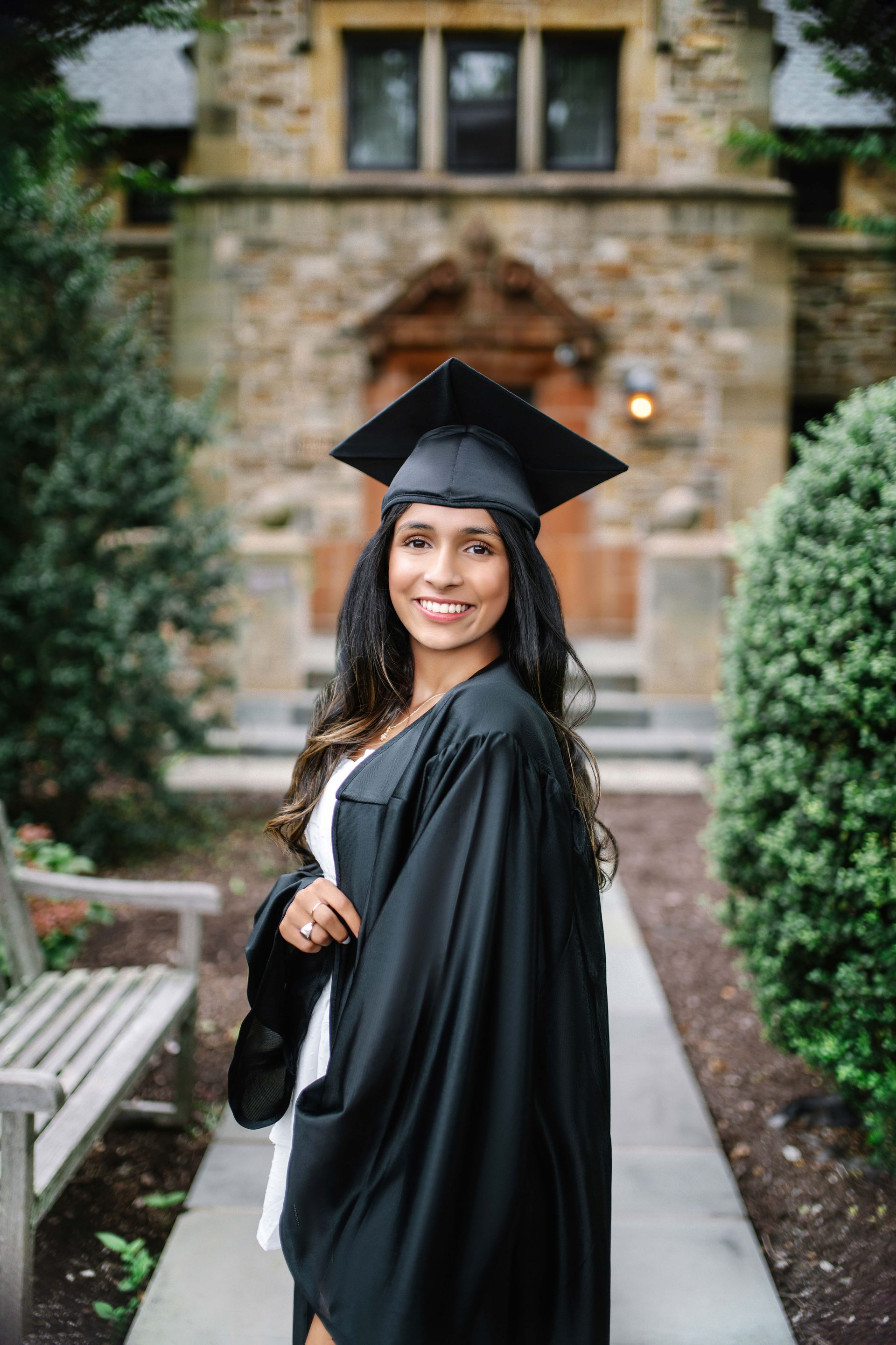 Woman in graduation cap and gown smiles in front of a brick building with a stone archway.