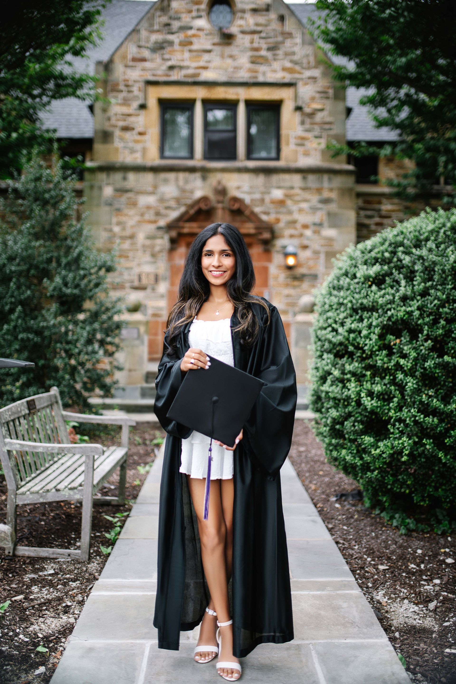 Young woman in graduation gown and cap smiles in front of a stone building, holding cap.