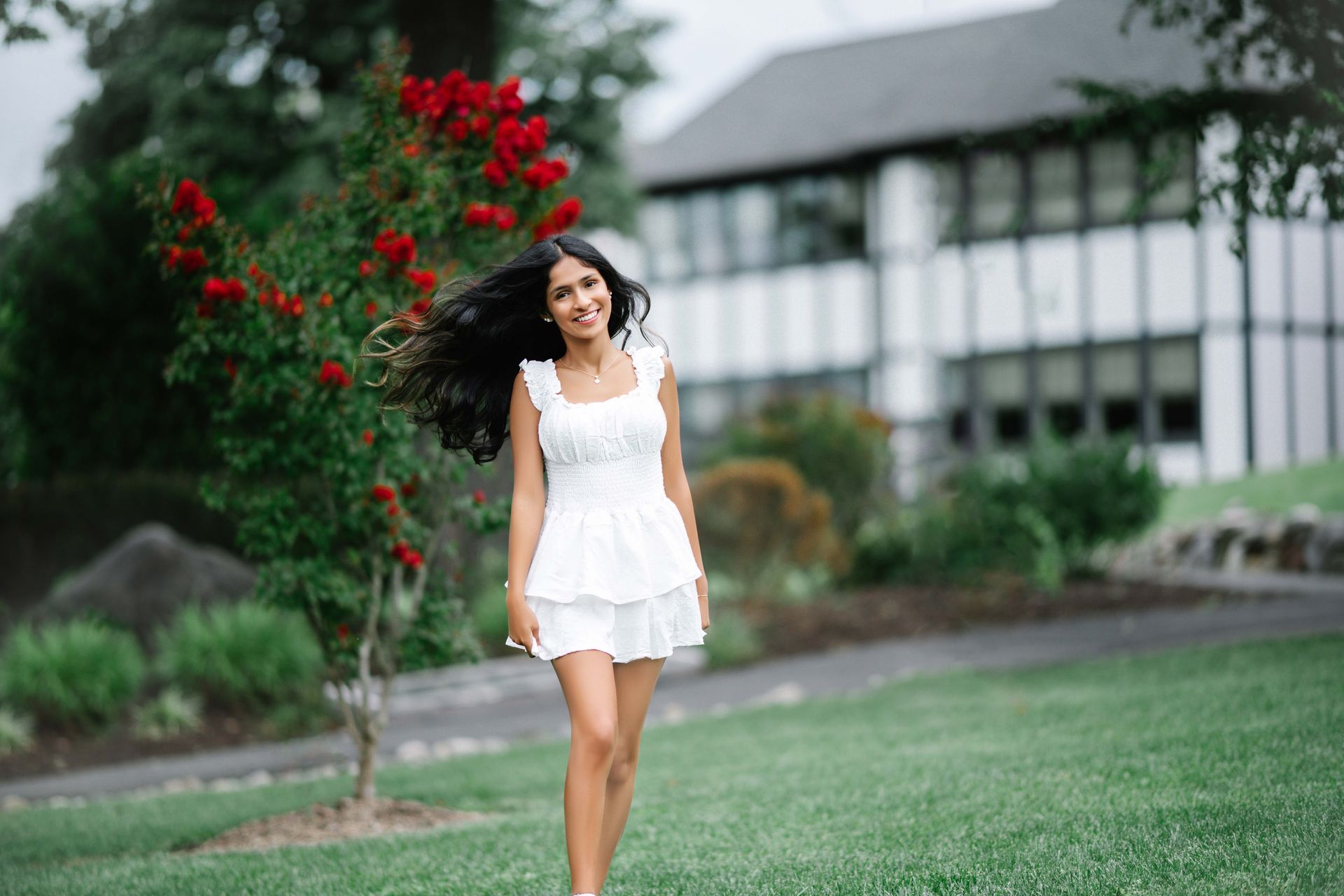 Woman in white dress smiles while walking outdoors in front of a building and tree with red flowers.