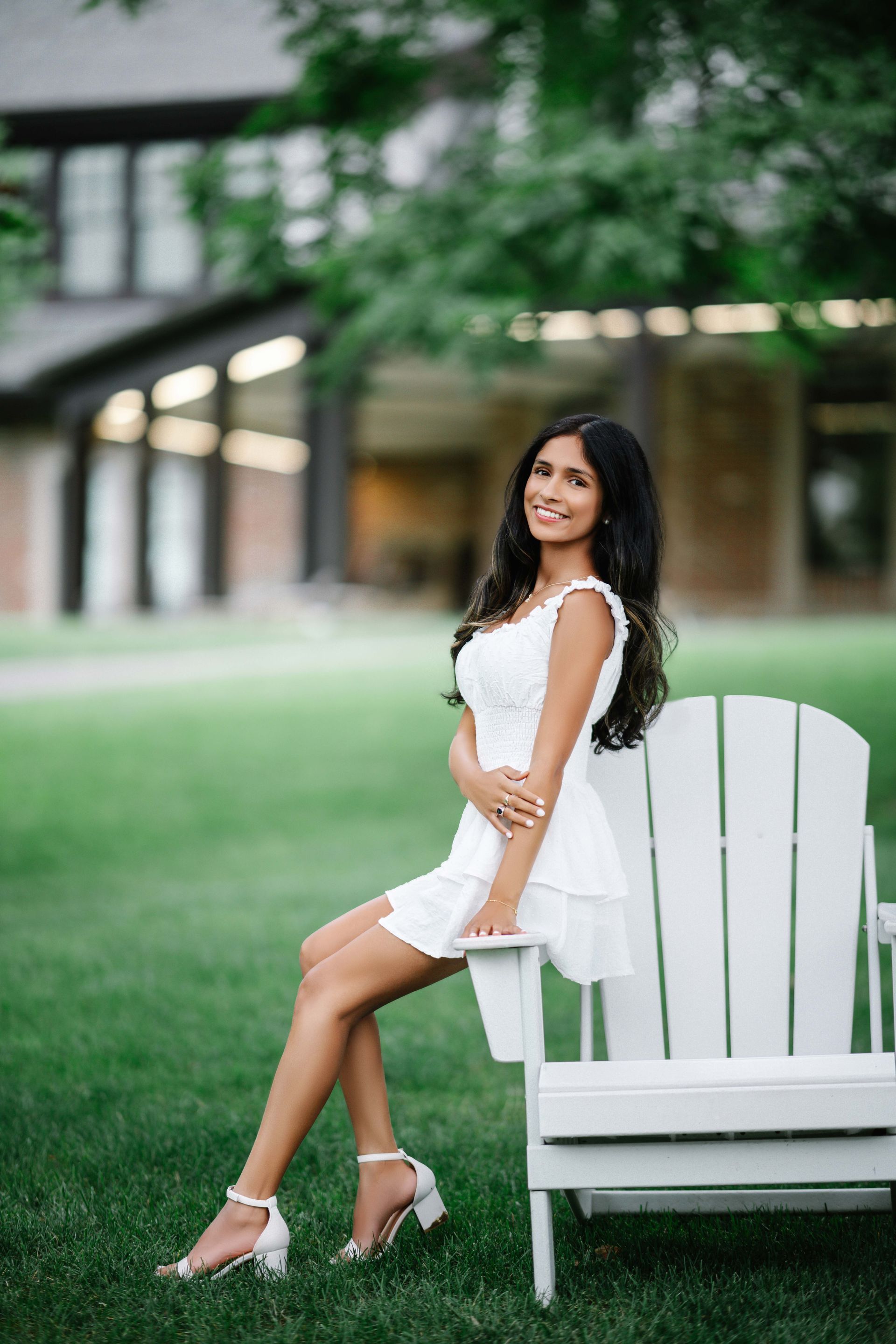 Woman in white dress smiles, seated on white chair in front of a building and green lawn.