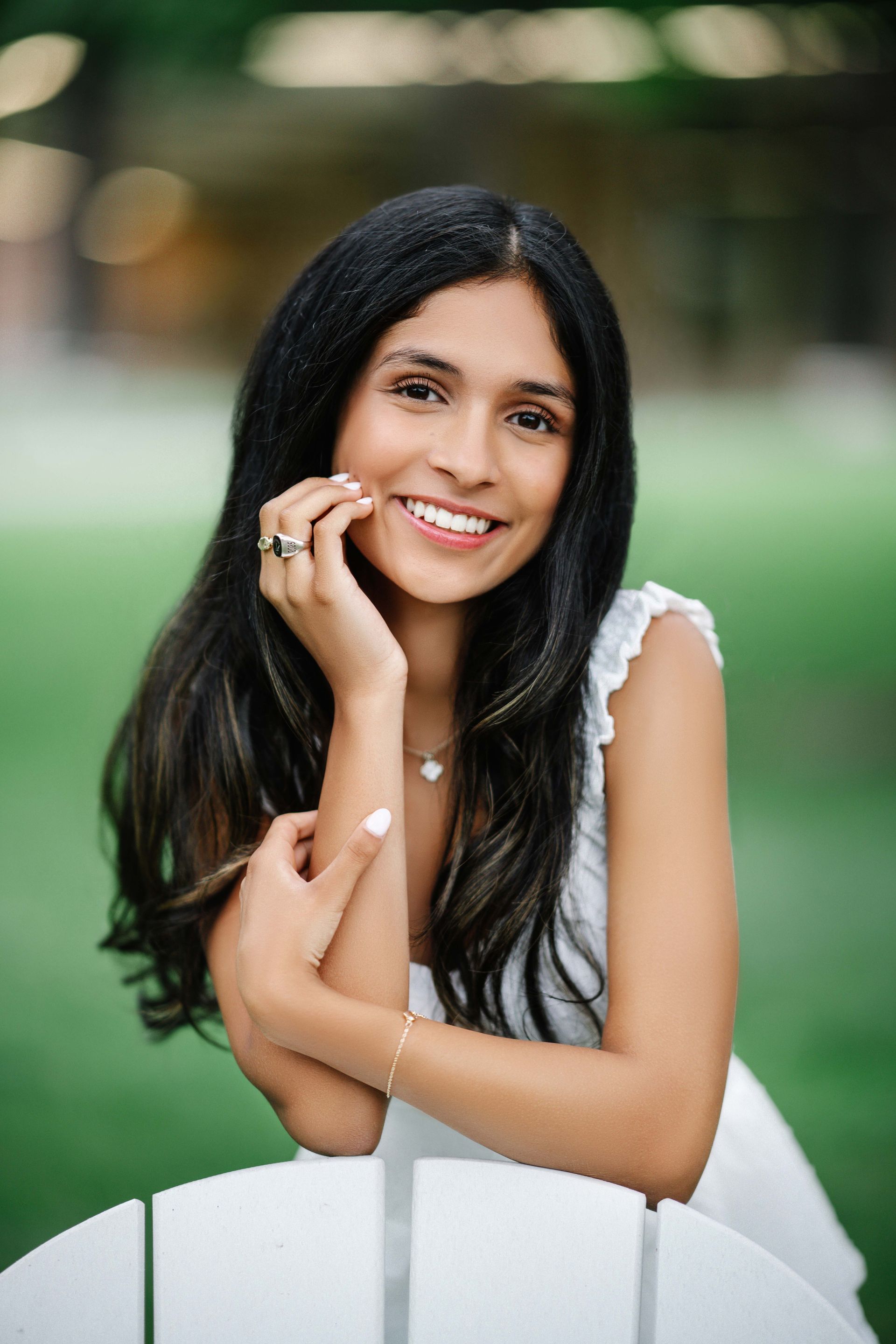 Woman with long dark hair smiles while leaning on a white chair in a park setting.
