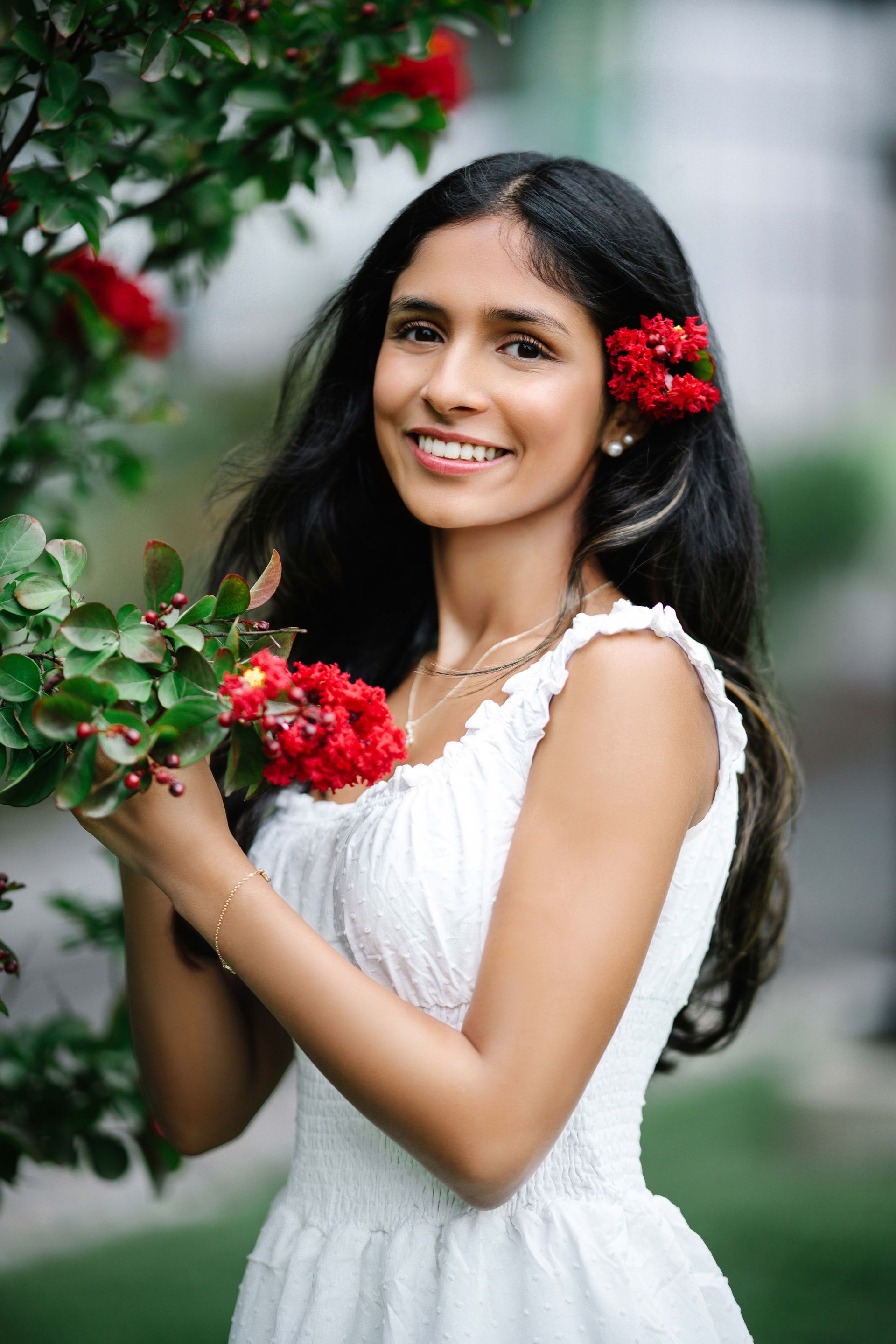 Woman in white dress with red flowers in hair and hand, smiling in garden.
