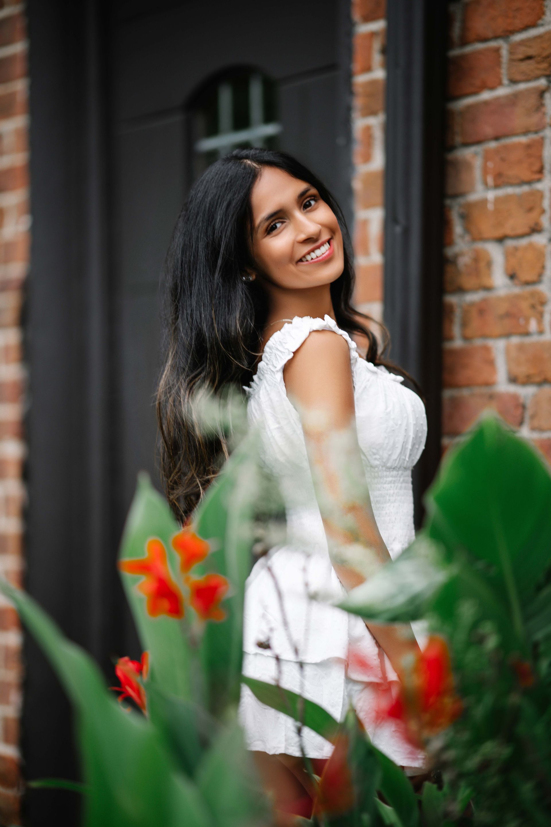 Woman in white dress smiling next to brick wall and red flowers.