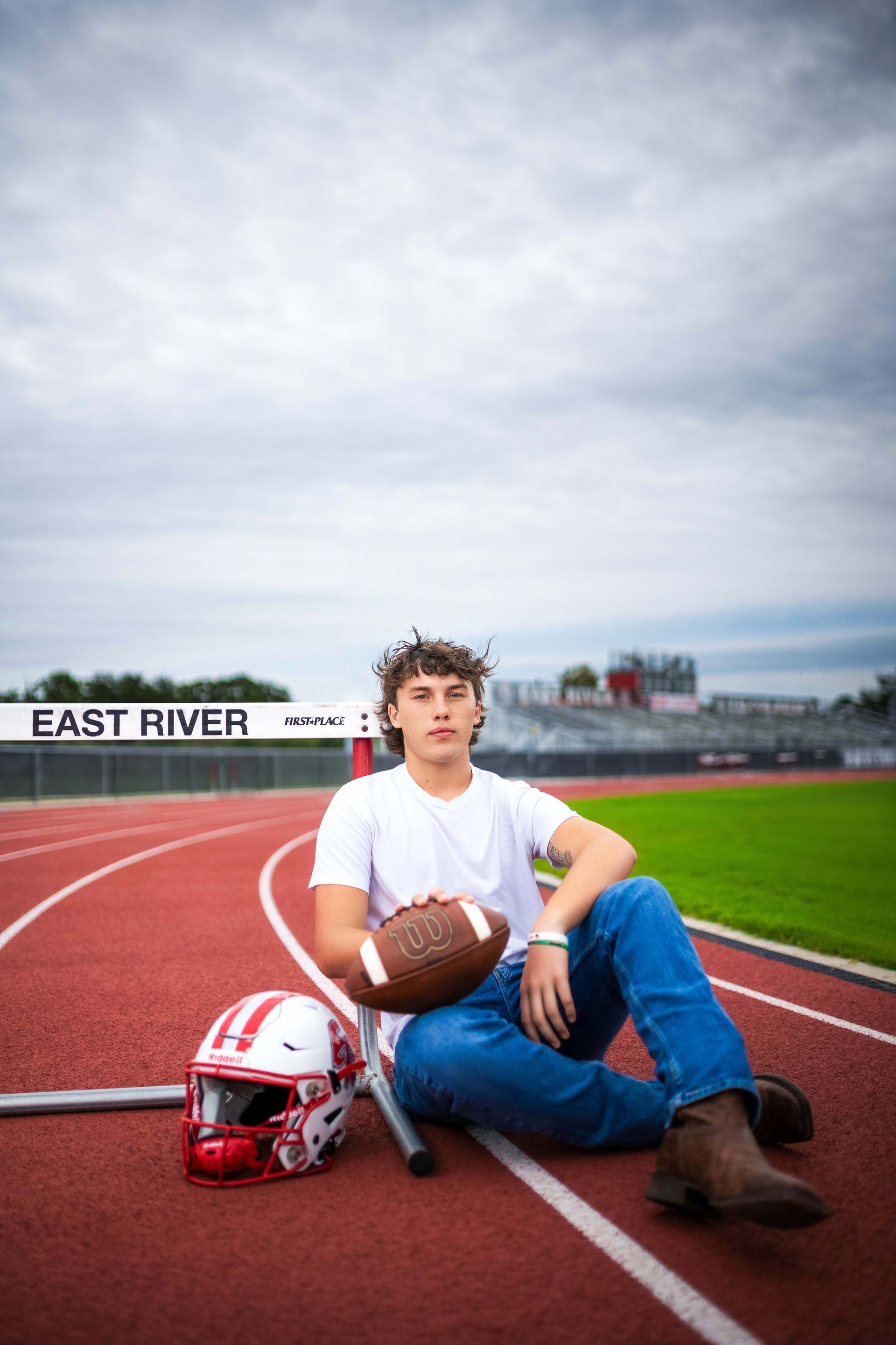 Young man sitting on a track, holding football. White shirt, blue jeans, boots. Football helmet to the side.
