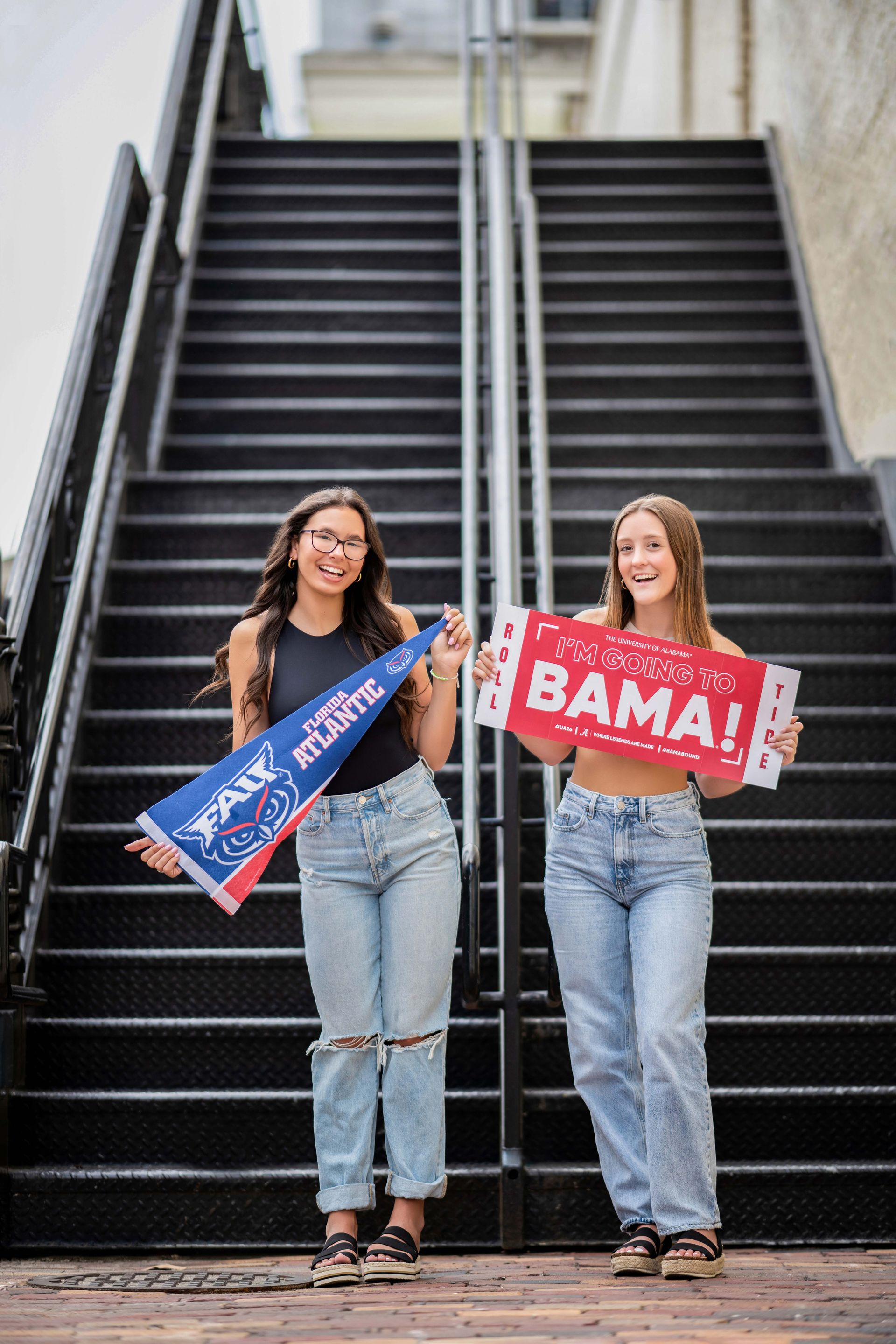 Two young women smiling, holding pennants on a staircase. One reads 