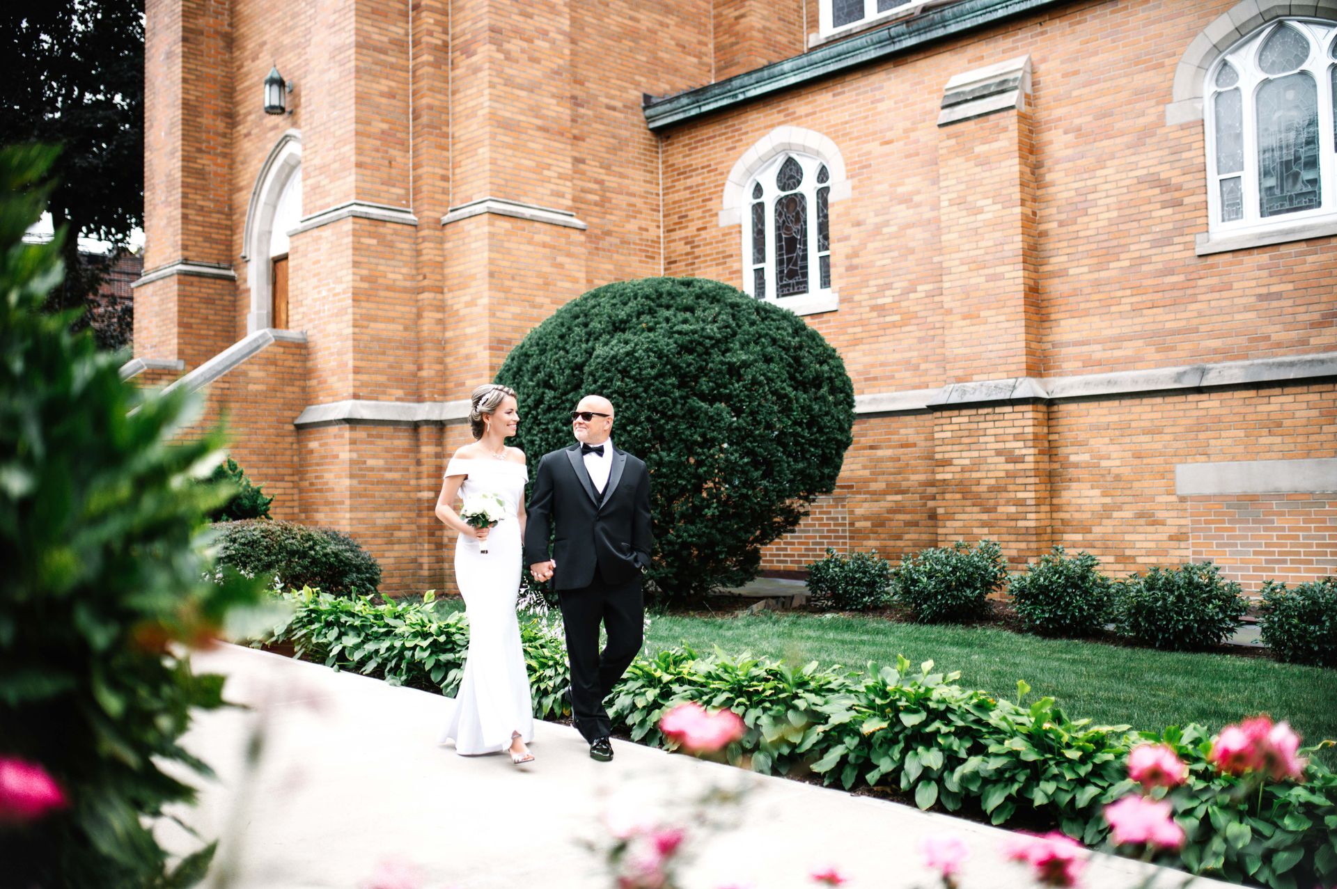 Bride and groom walk hand-in-hand in front of a brick church; she wears white, he wears a tux.