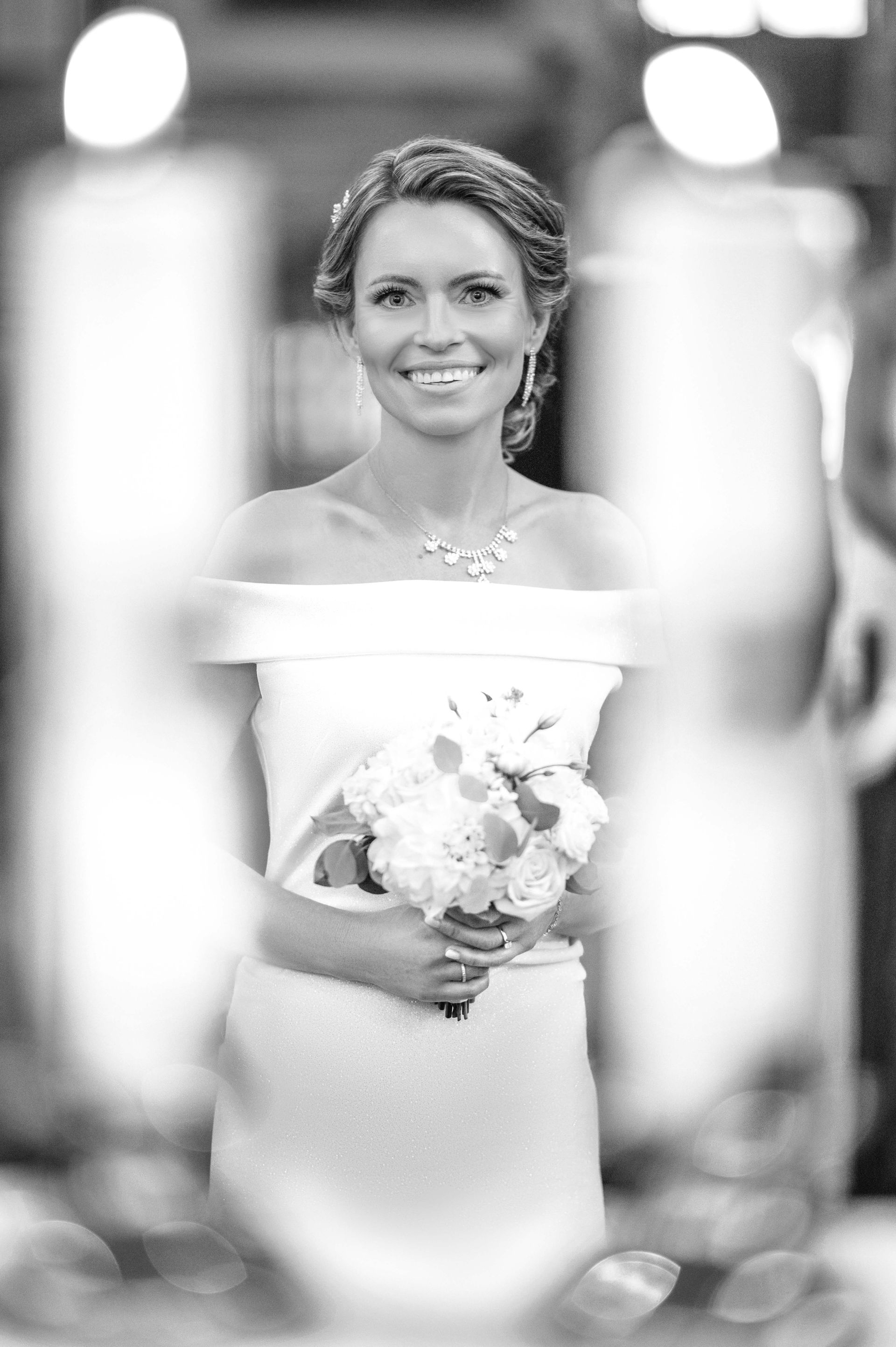 Bride in off-the-shoulder gown holding flowers, smiling. Black and white photo, blurred foreground.