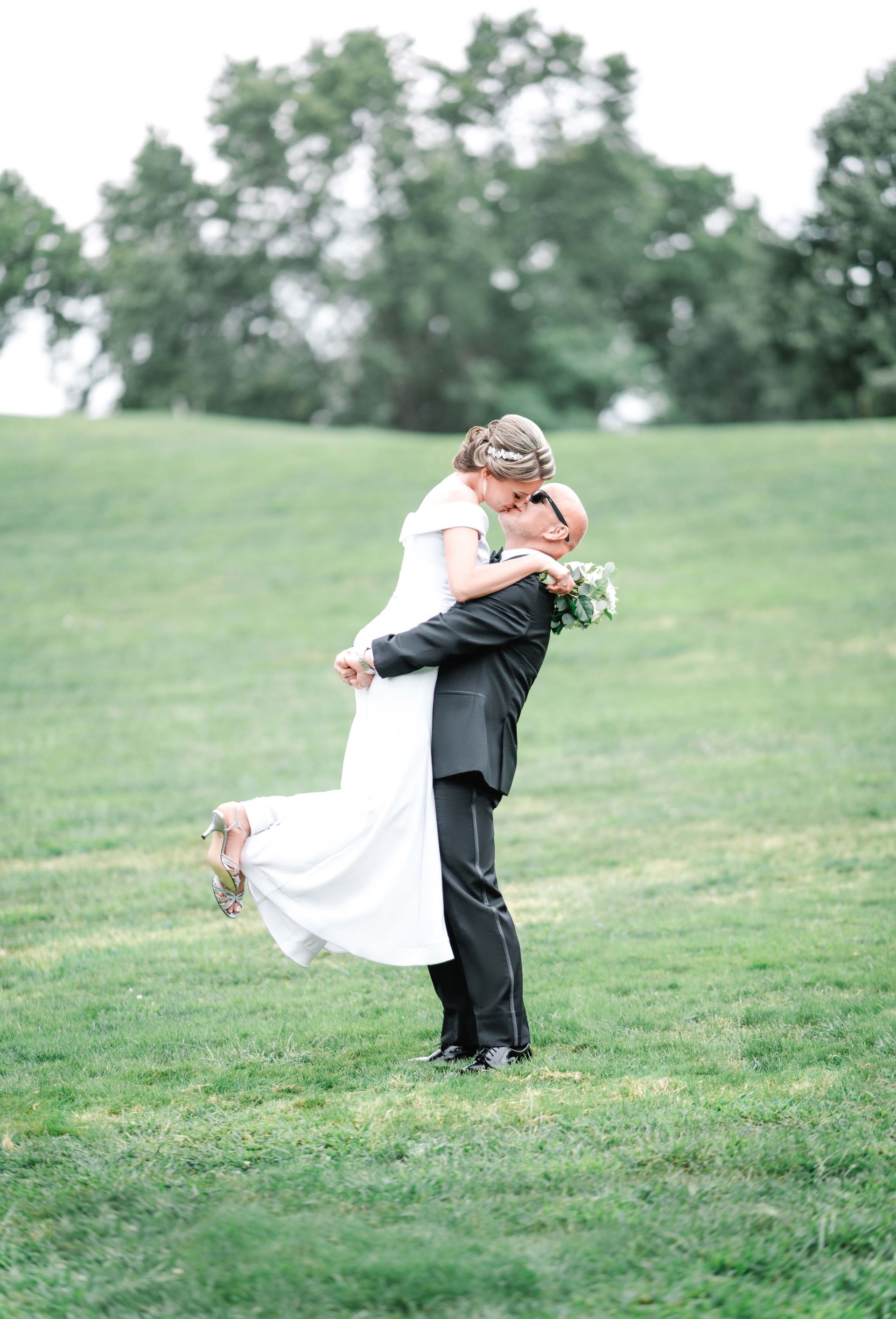 Groom lifting bride, kissing. They stand in a grassy field, trees in the background.