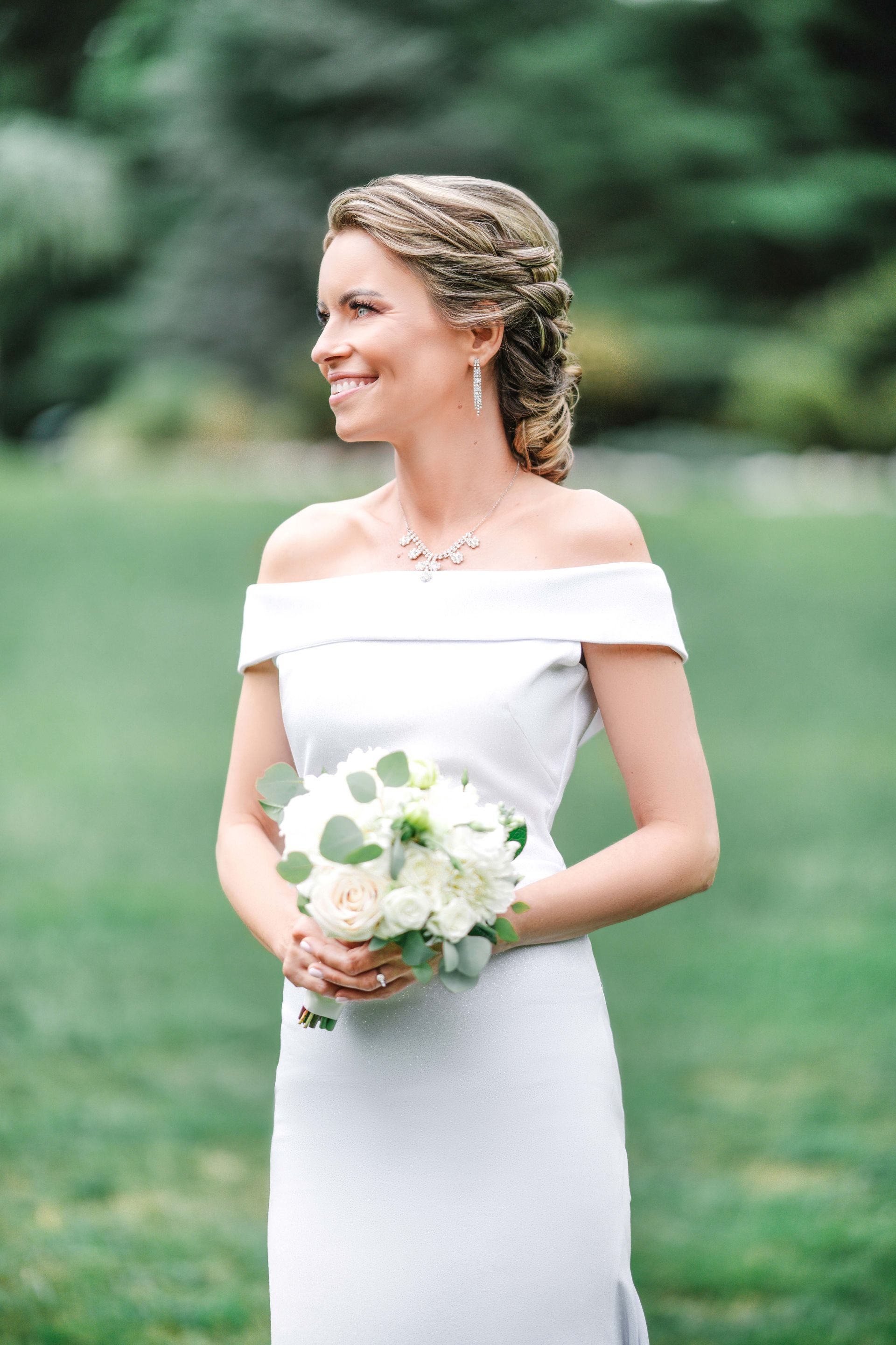 Bride in white off-shoulder dress, smiling, holding bouquet, looking left, outdoor setting.