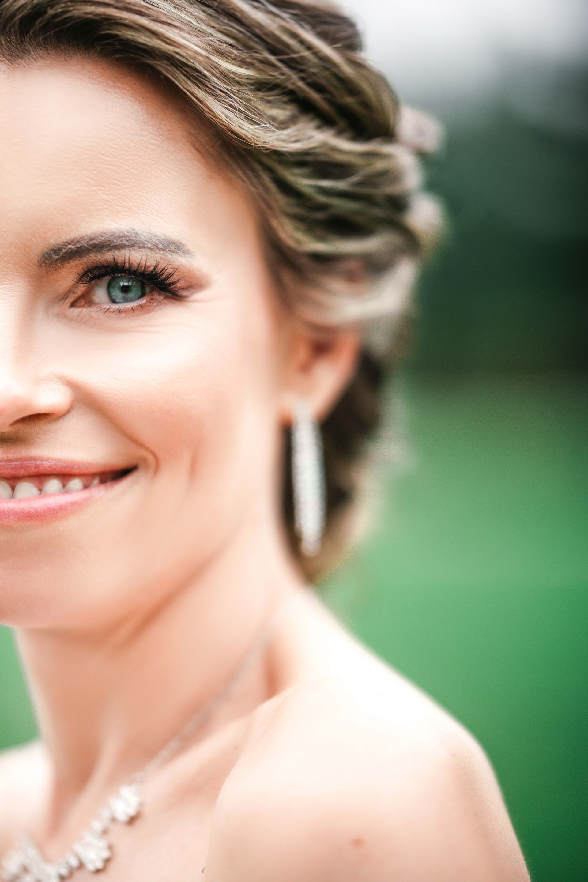 Woman with blue eyes smiles, wearing a necklace and earrings, outdoors.