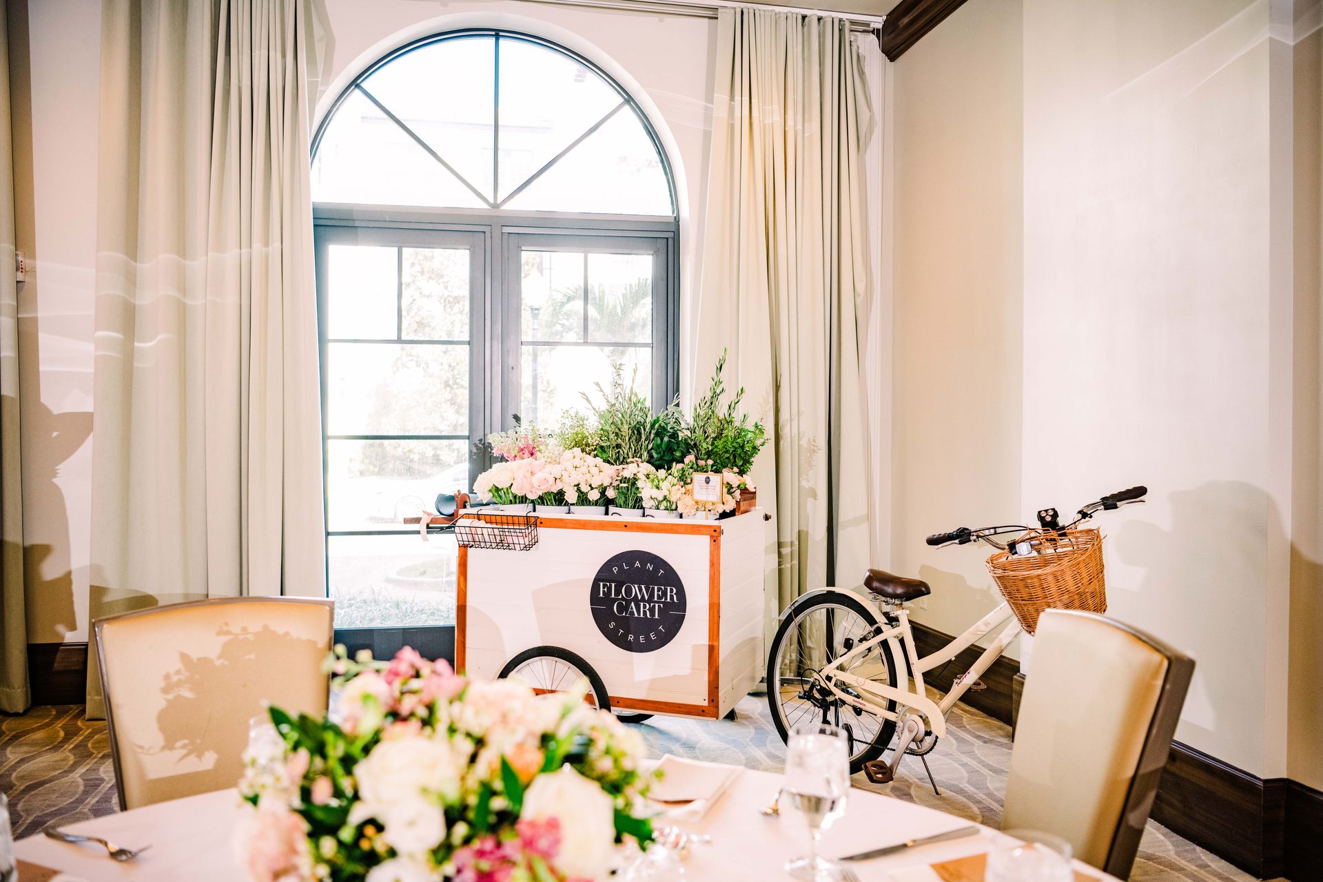 Wedding reception setup with flower cart and bicycle, near a window.