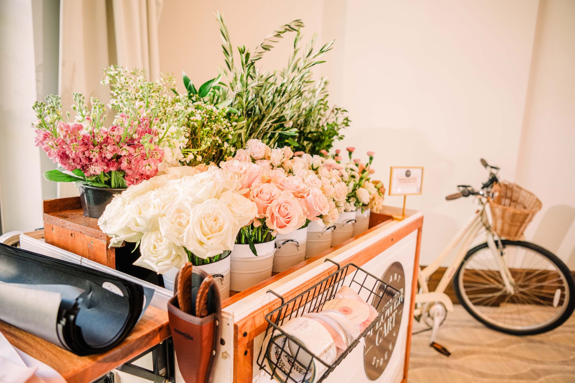 Flower cart with assorted blooms, a bike, and small sign in a bright setting.