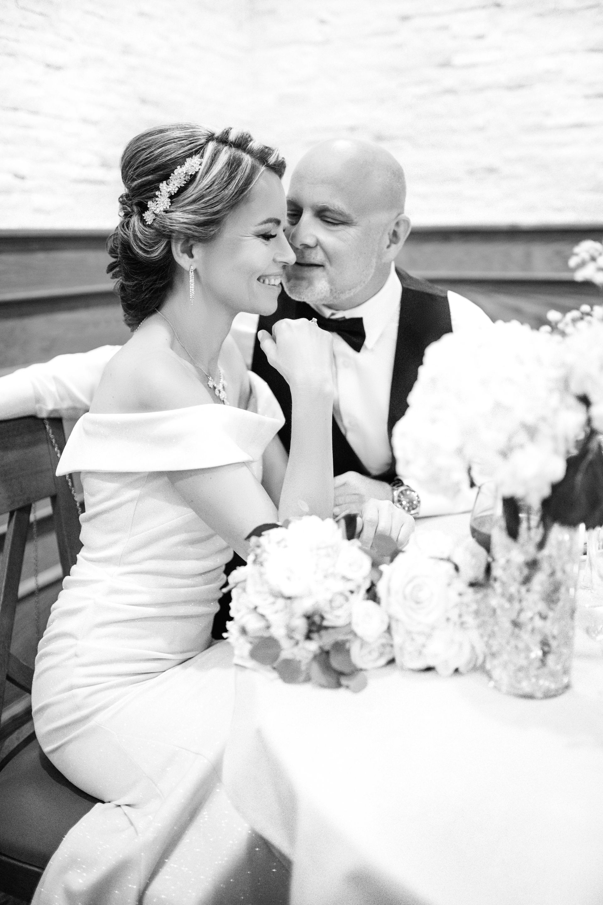 Bride and groom embrace at a table, black and white. Bride in off-shoulder dress, hair up with headpiece. Groom in suit, bow tie.