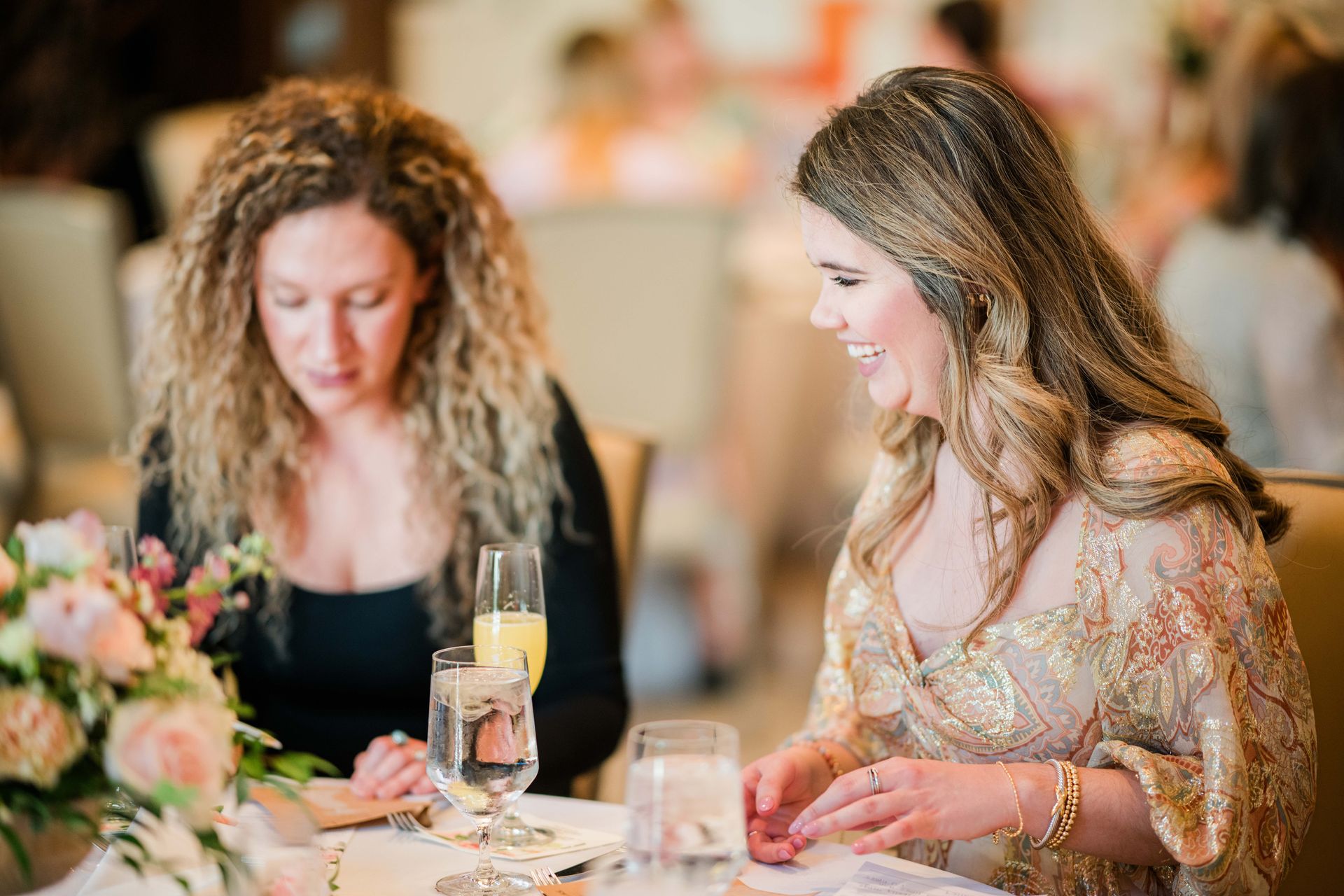 Two women at a table, one smiling in a sparkly gold top, the other with curly hair.  Blurred background of a restaurant.