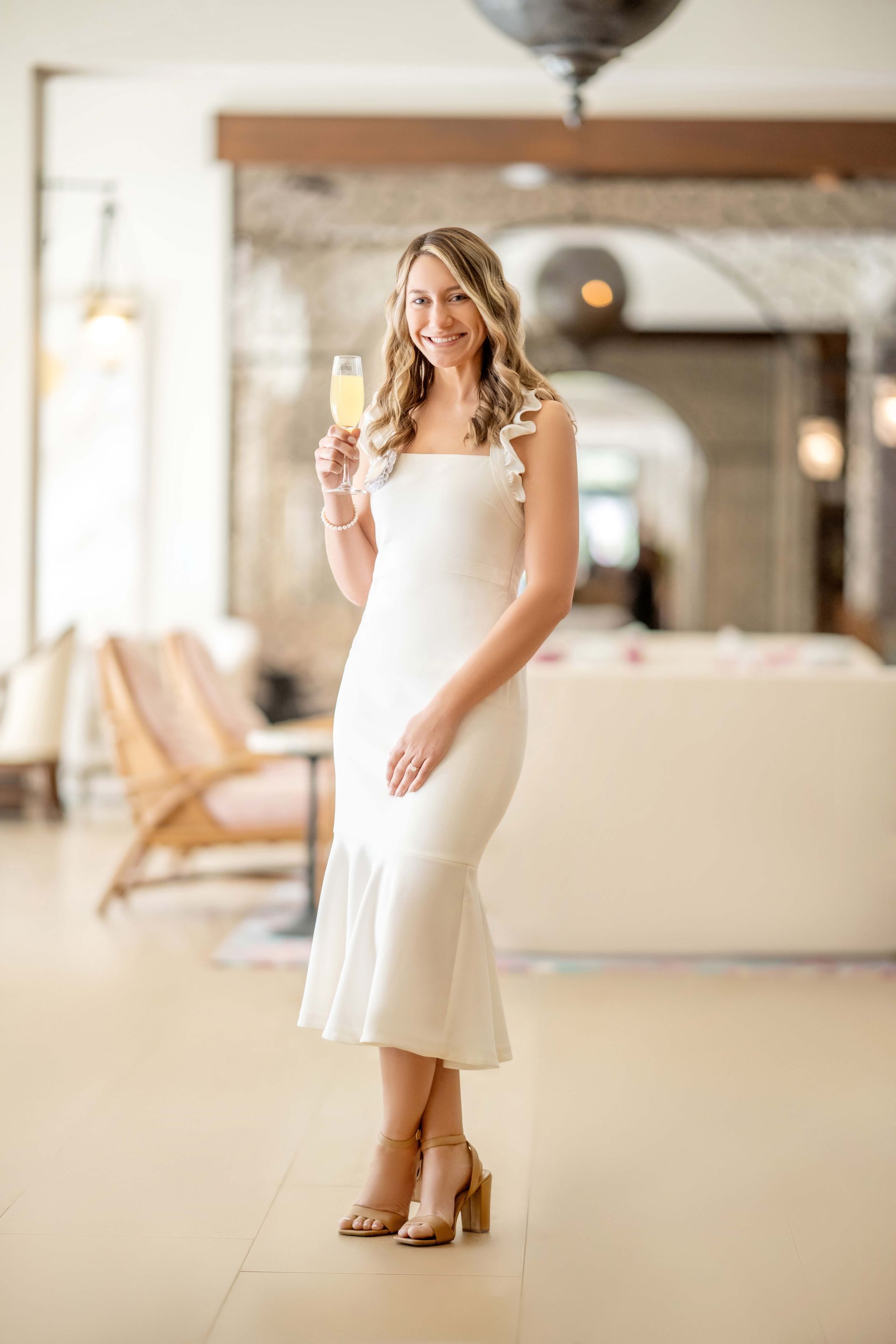 Woman in white dress, holding drink, smiles. Standing in light-filled room with high ceiling.