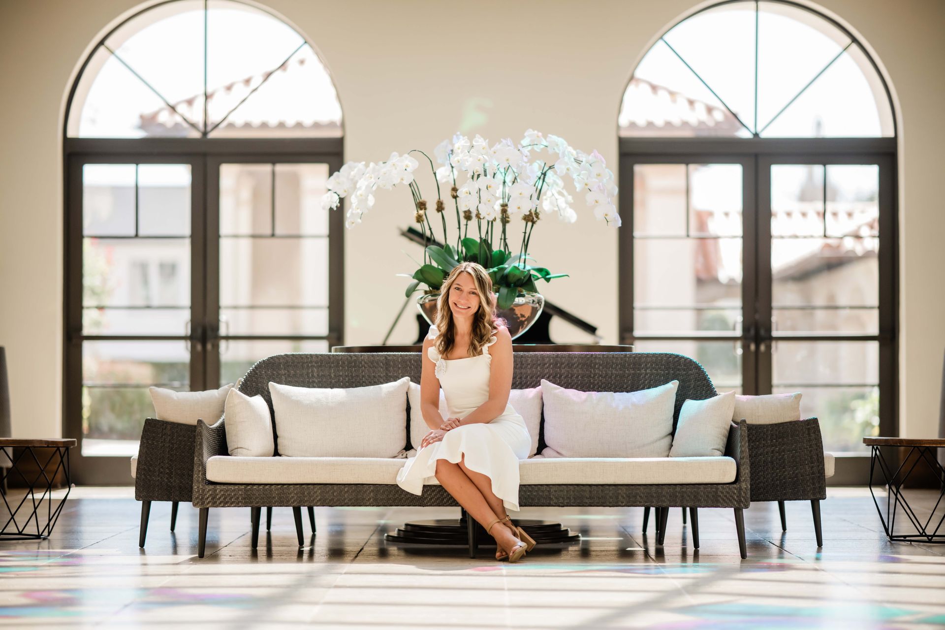Woman in white dress sits on a couch in a bright lobby with large windows and large white flower arrangement.