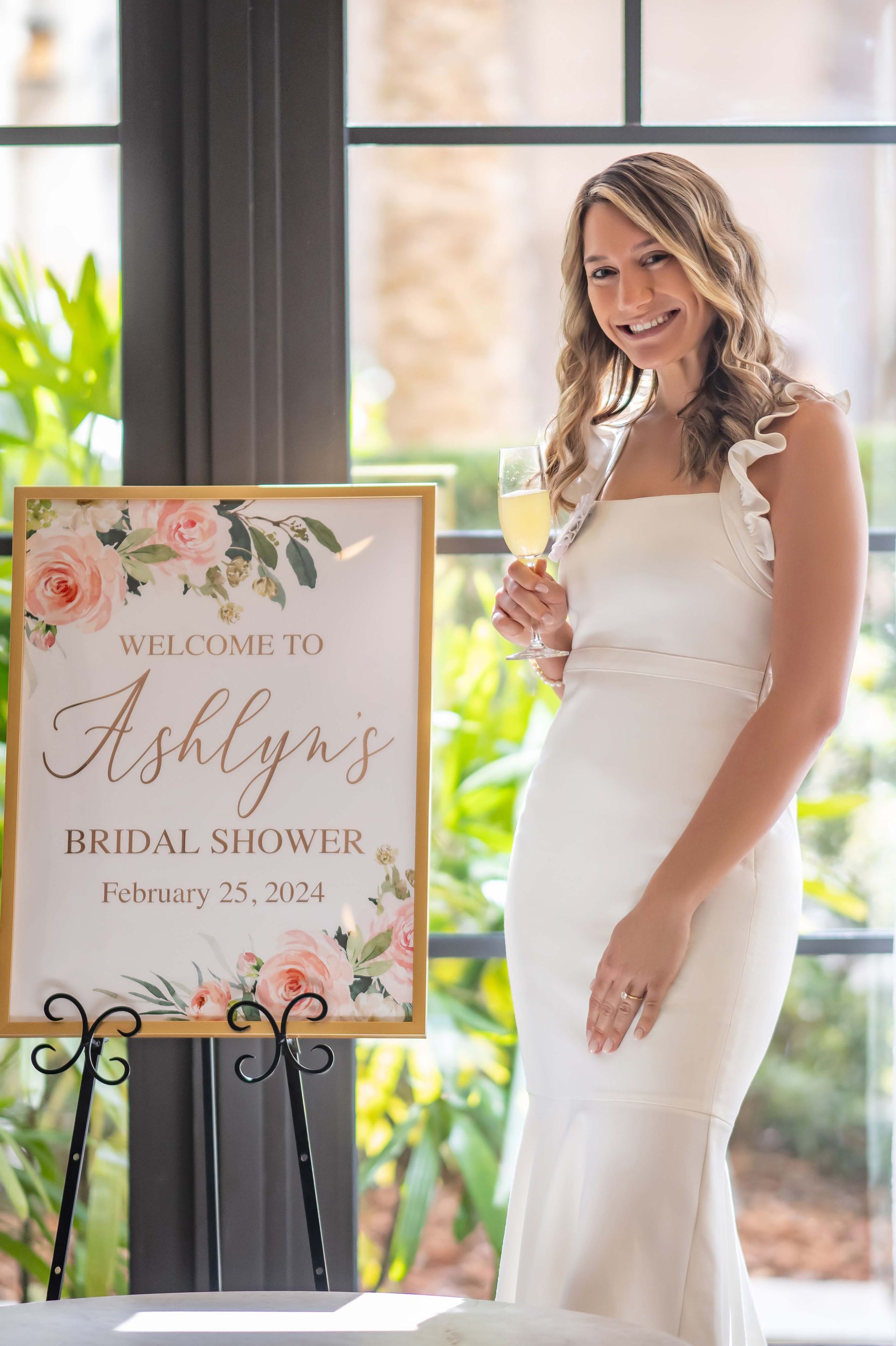 Woman in white dress holding champagne at Ashlyn's bridal shower. Sign says 