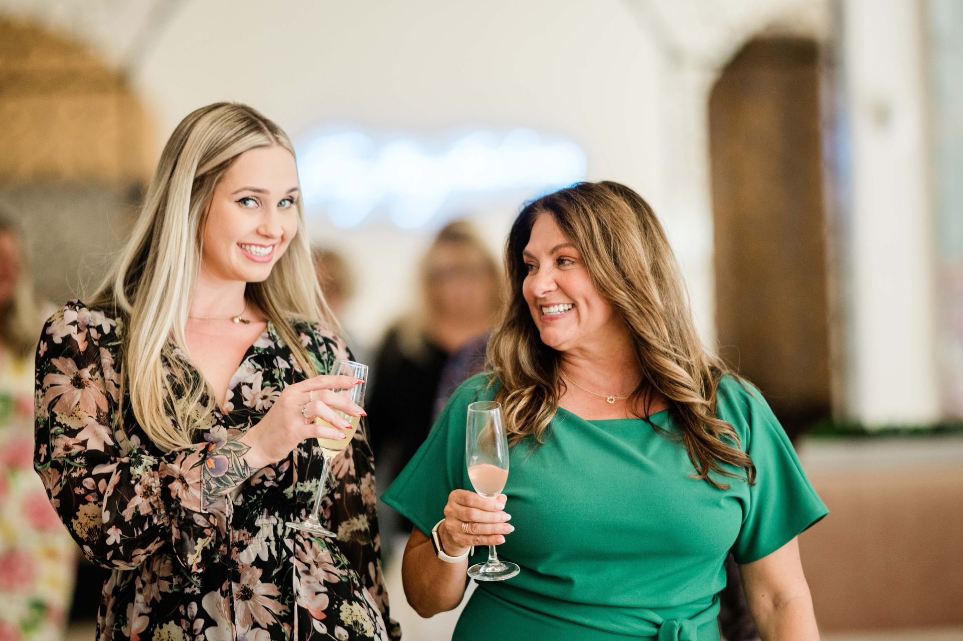Two smiling women, one blonde, one brunette, holding champagne glasses in an elegant setting.