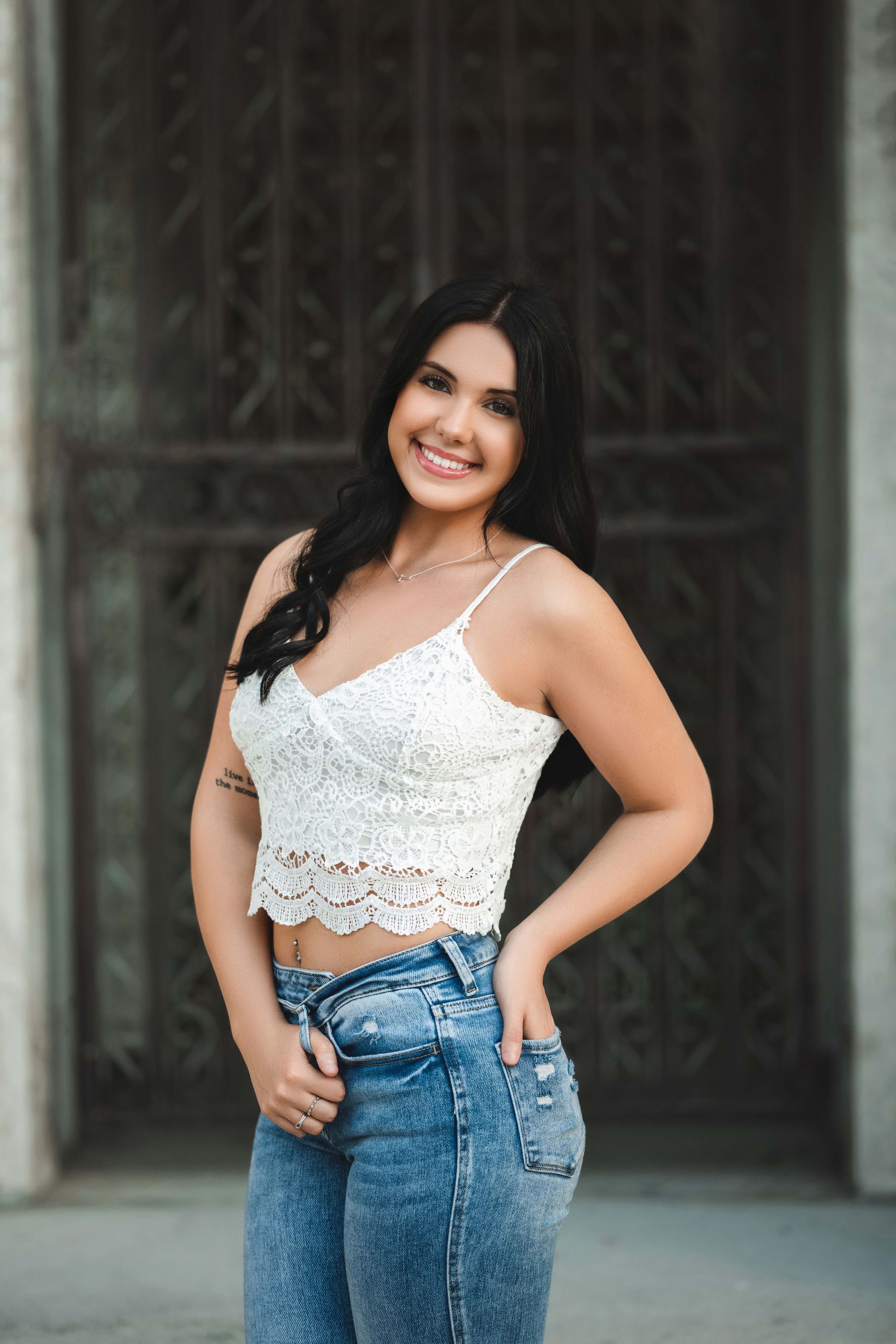 Woman with dark hair smiles, wearing white lace top and acid wash jeans, standing in front of a dark gate.