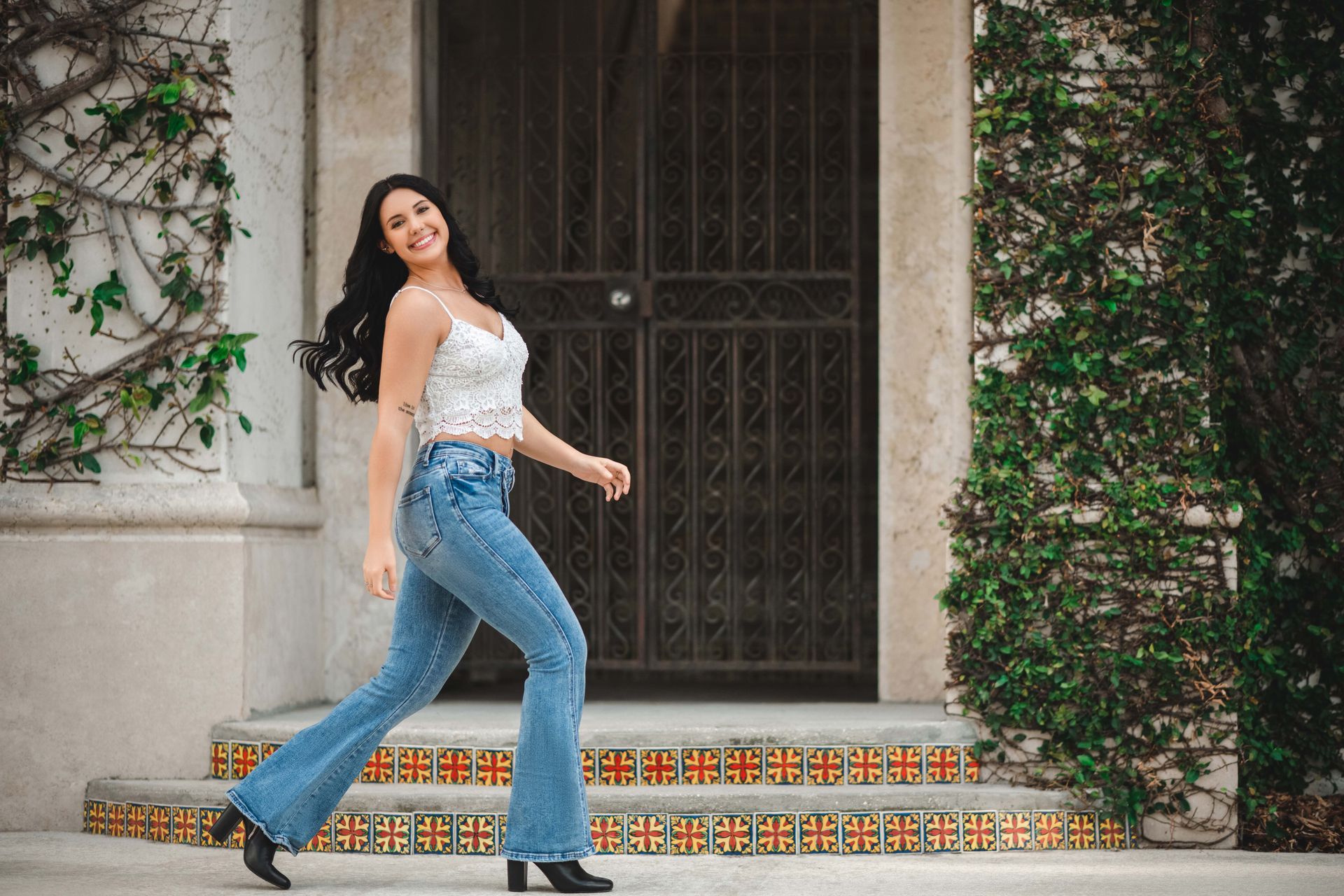 Woman in white top and flared jeans smiles, walking near a building entrance with a gate.