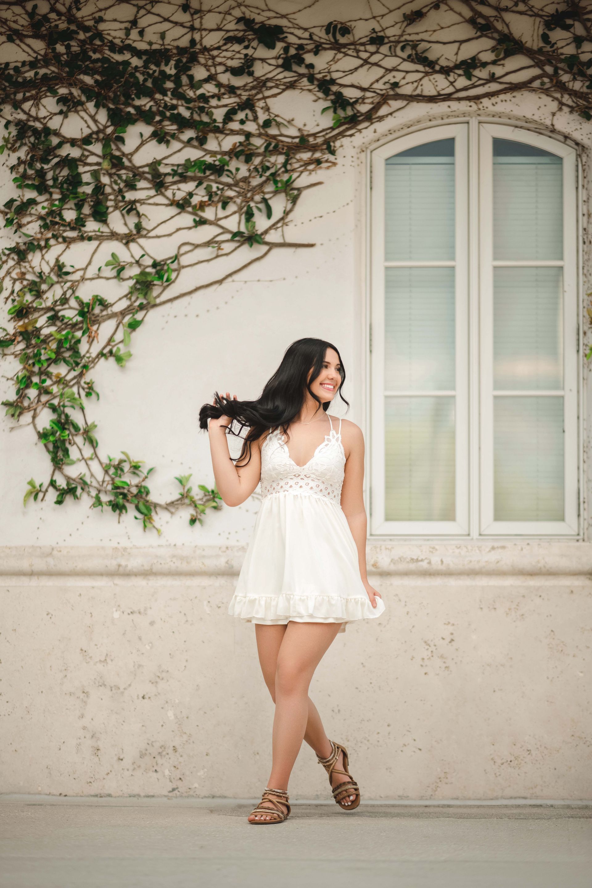 Woman in white dress poses near ivy-covered wall and window.