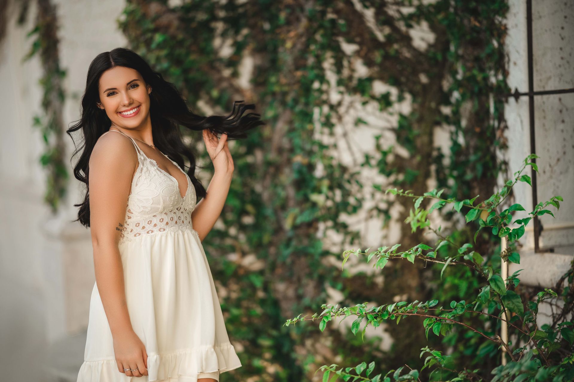 Woman in white dress, smiling, near ivy-covered wall, touching hair.