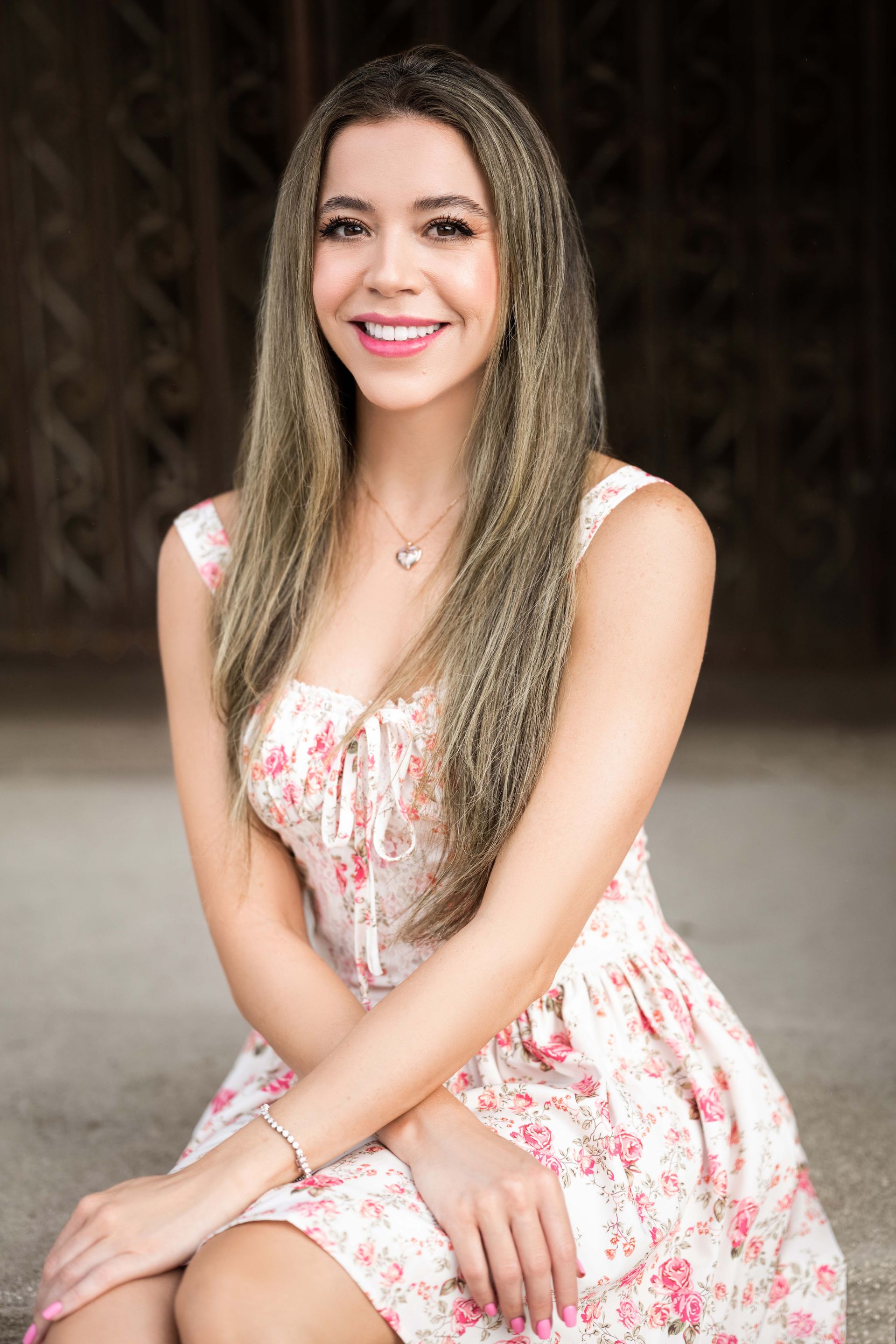 Woman with long blonde hair smiles in a floral dress, seated outdoors.