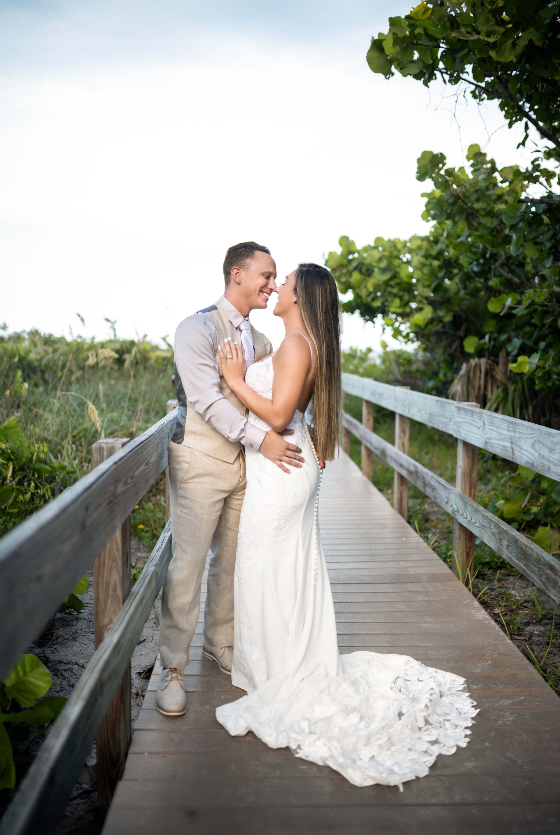 Groom in beige suit embraces bride in white gown on a wooden bridge. They smile, with ocean and greenery behind them.
