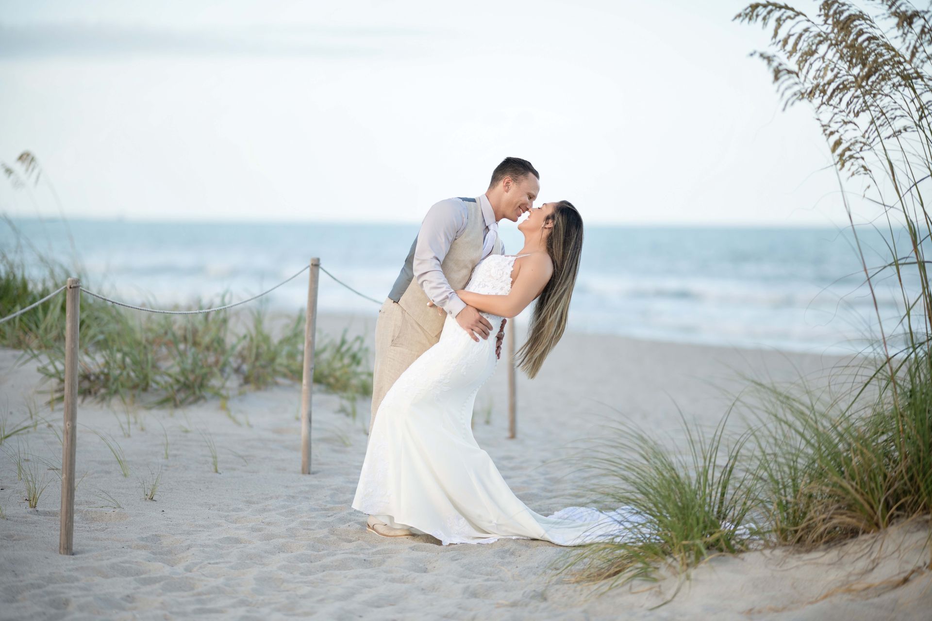 Couple leans in to kiss on a sandy beach; woman wears a white gown, man in a vest.
