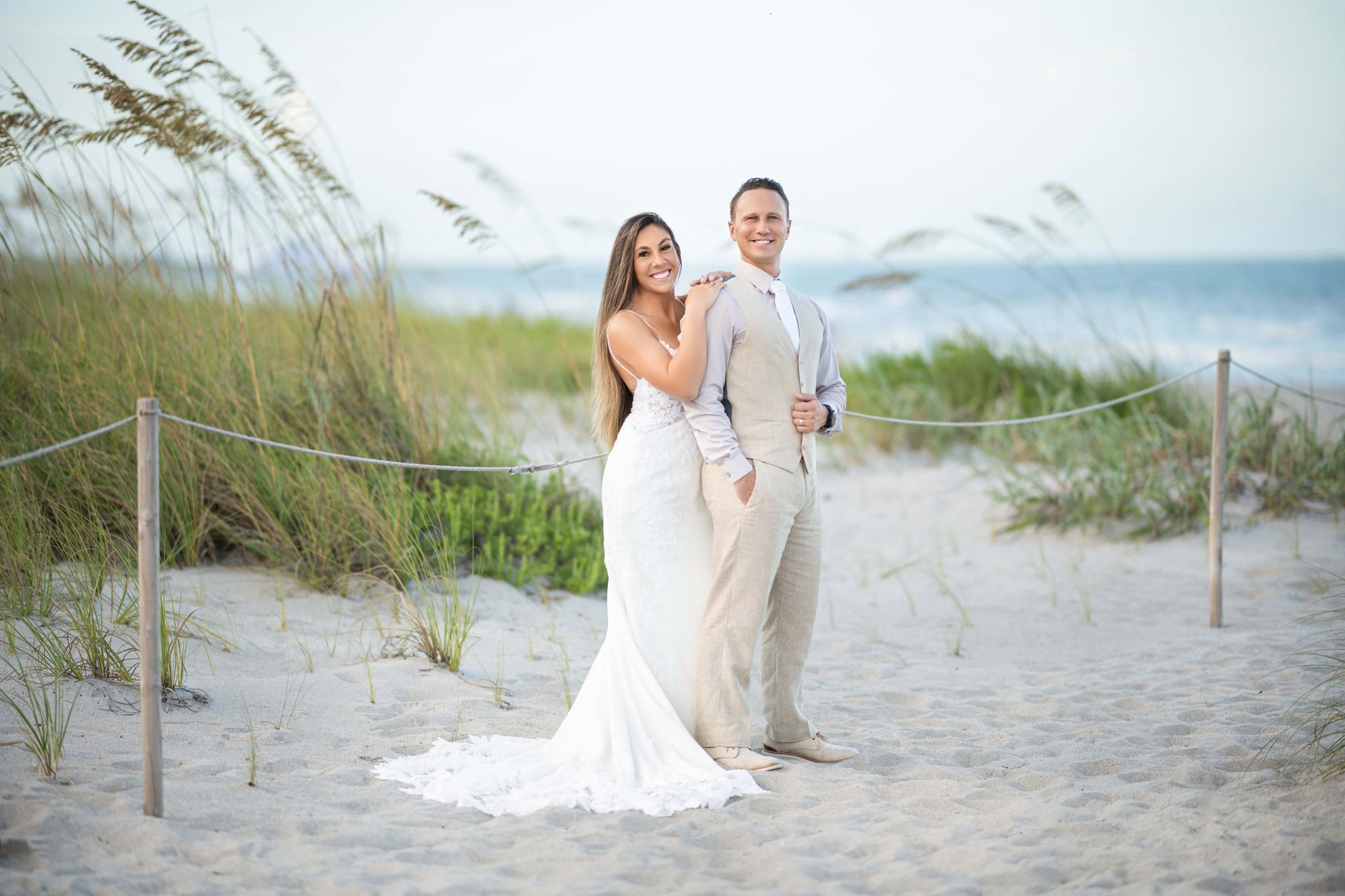 A bride and groom pose on a beach, the bride's arm on the groom's shoulder, with the ocean in the background.
