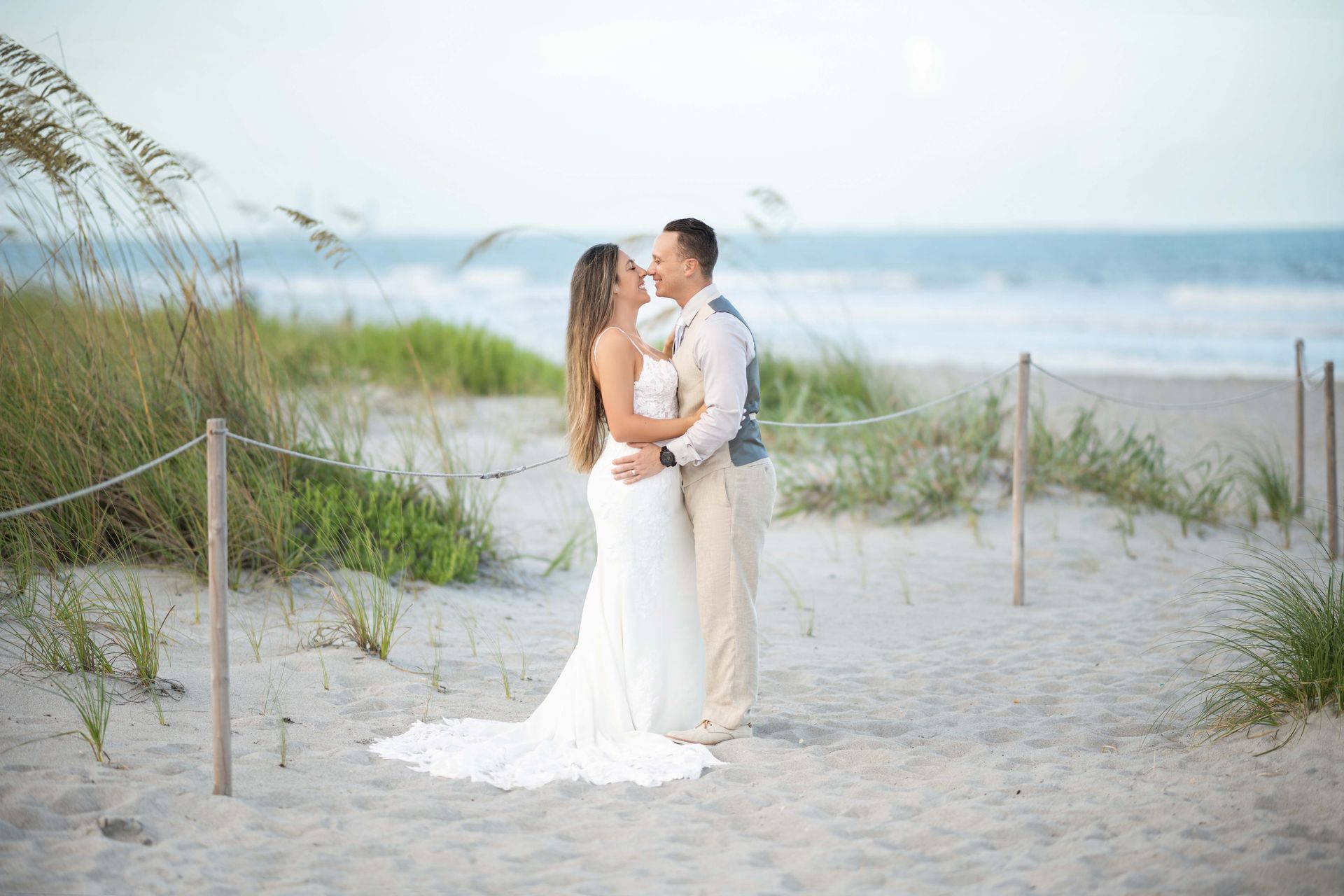 Couple embraces on a beach at sunset. The bride wears a white gown, and the groom wears a vest and khakis.