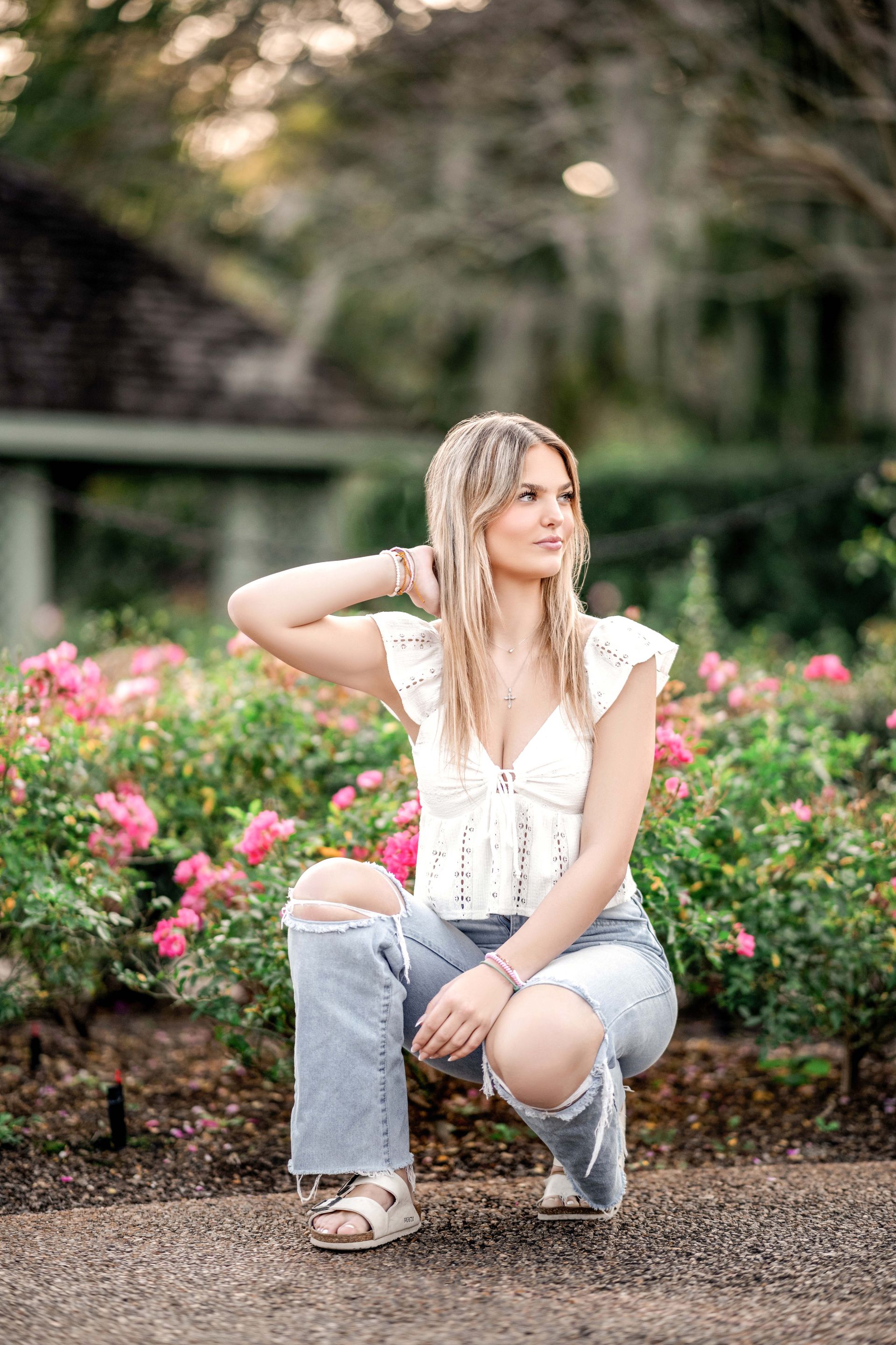 Blonde woman squats near pink flowers, wearing a white top and ripped jeans, looking away, in a garden.