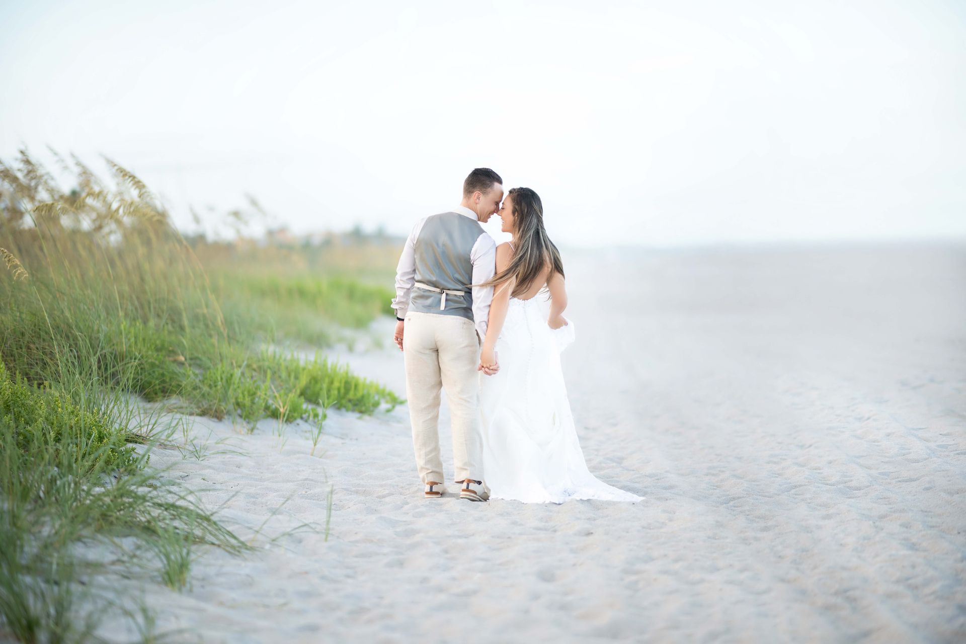 Bride and groom on a beach, holding hands and kissing. Bride in white gown, groom in vest and khaki pants.