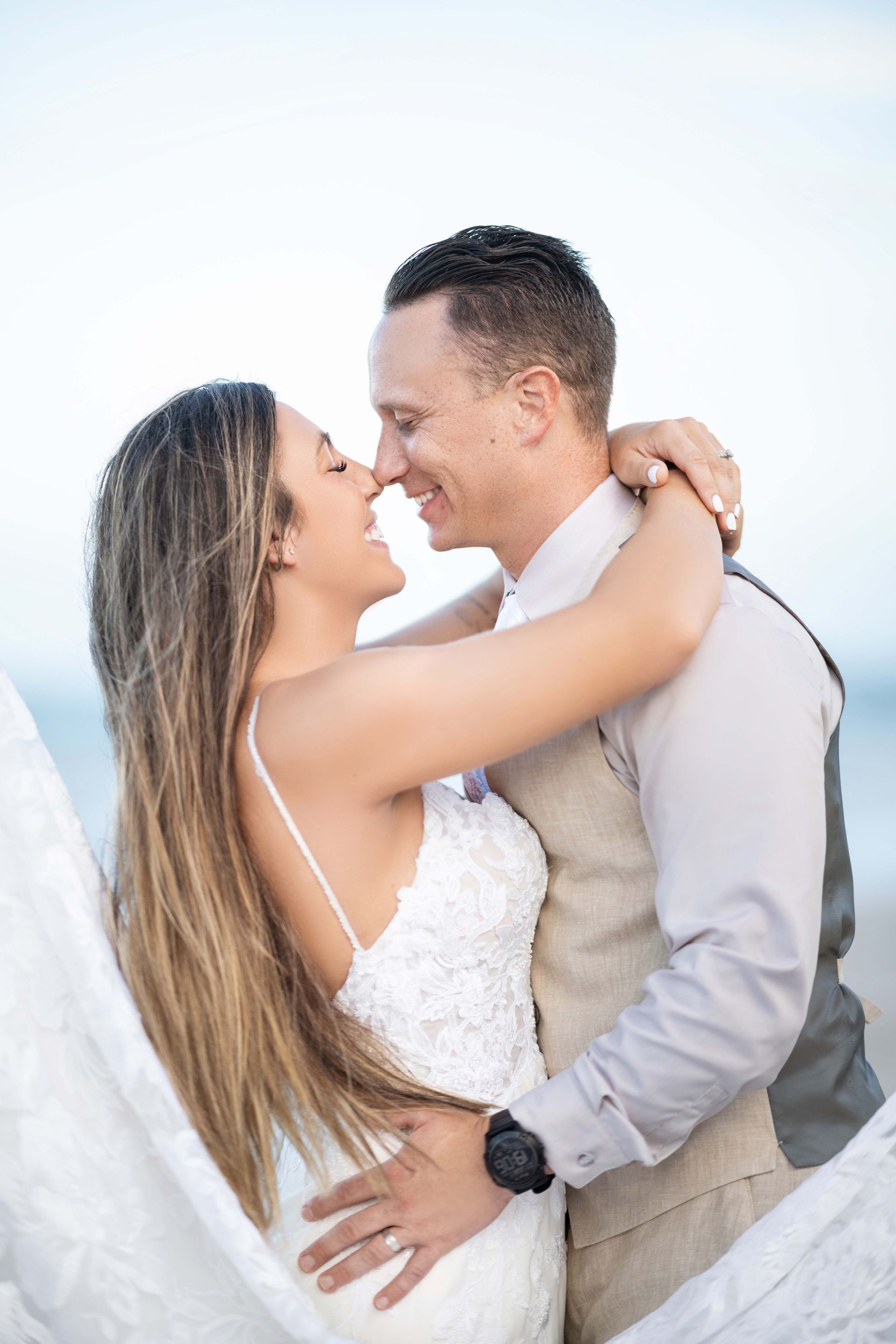 Bride and groom embracing on a beach; the bride smiles, the groom is gazing at her, blue ocean background.