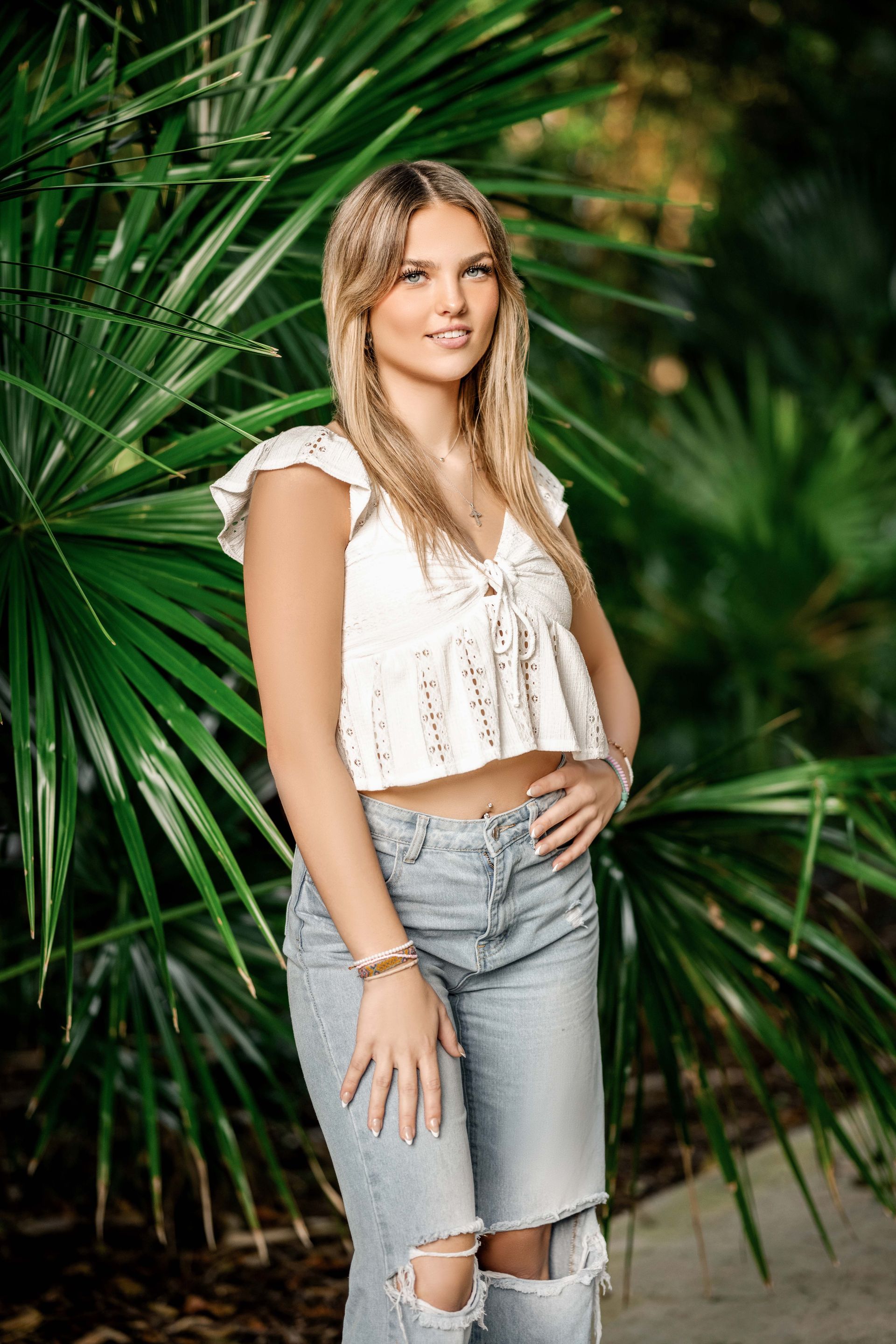 Young woman in white top and ripped jeans poses in front of green foliage.