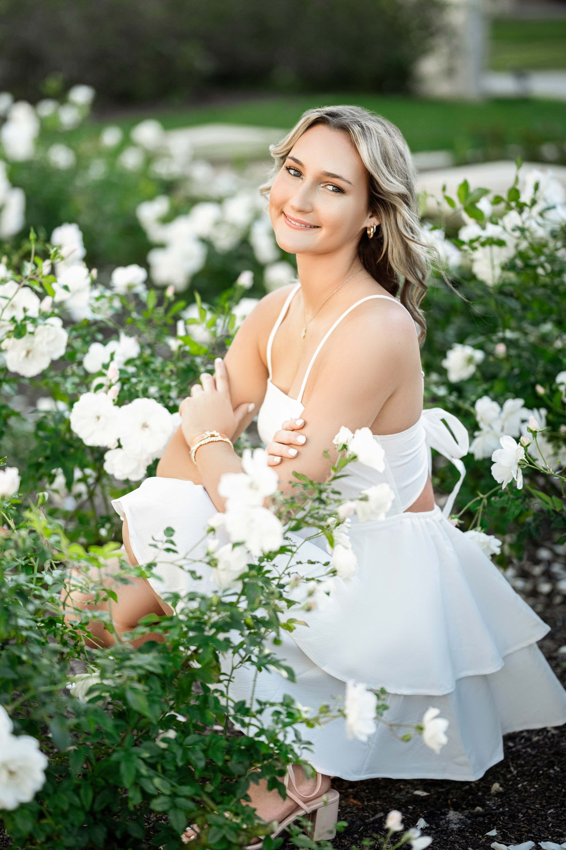 Blonde woman in white outfit smiles amidst white roses in garden.
