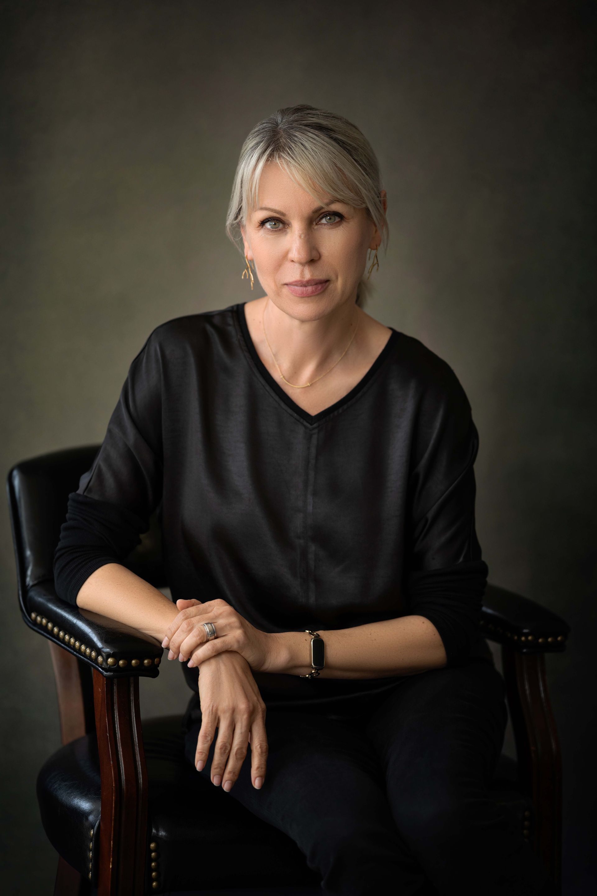 Woman with silver hair in a black shirt and pants, seated in a dark chair, looking forward.